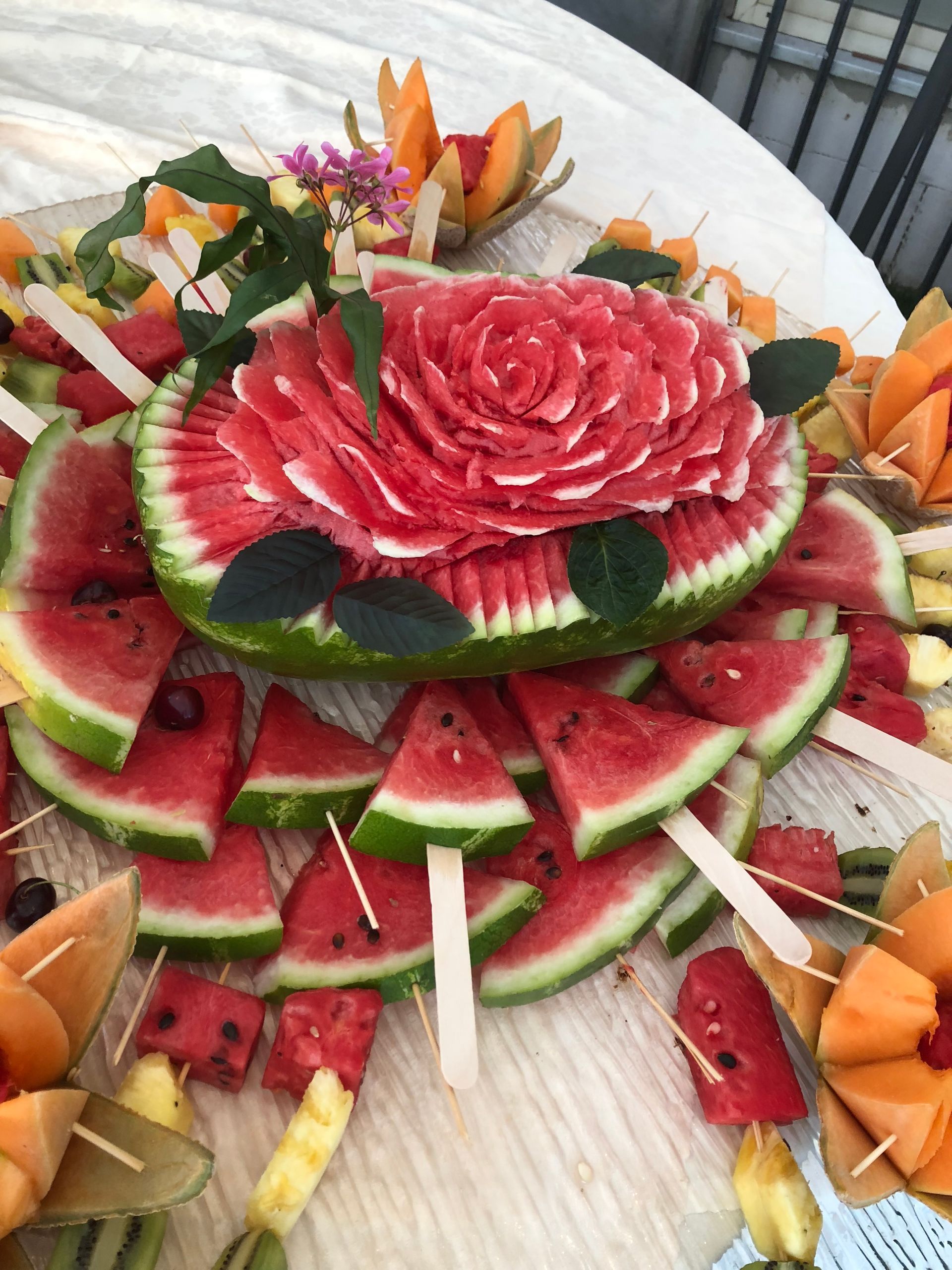 Centrotavola intagliato a forma di anguria con fette di frutta e spiedini su una superficie bianca.