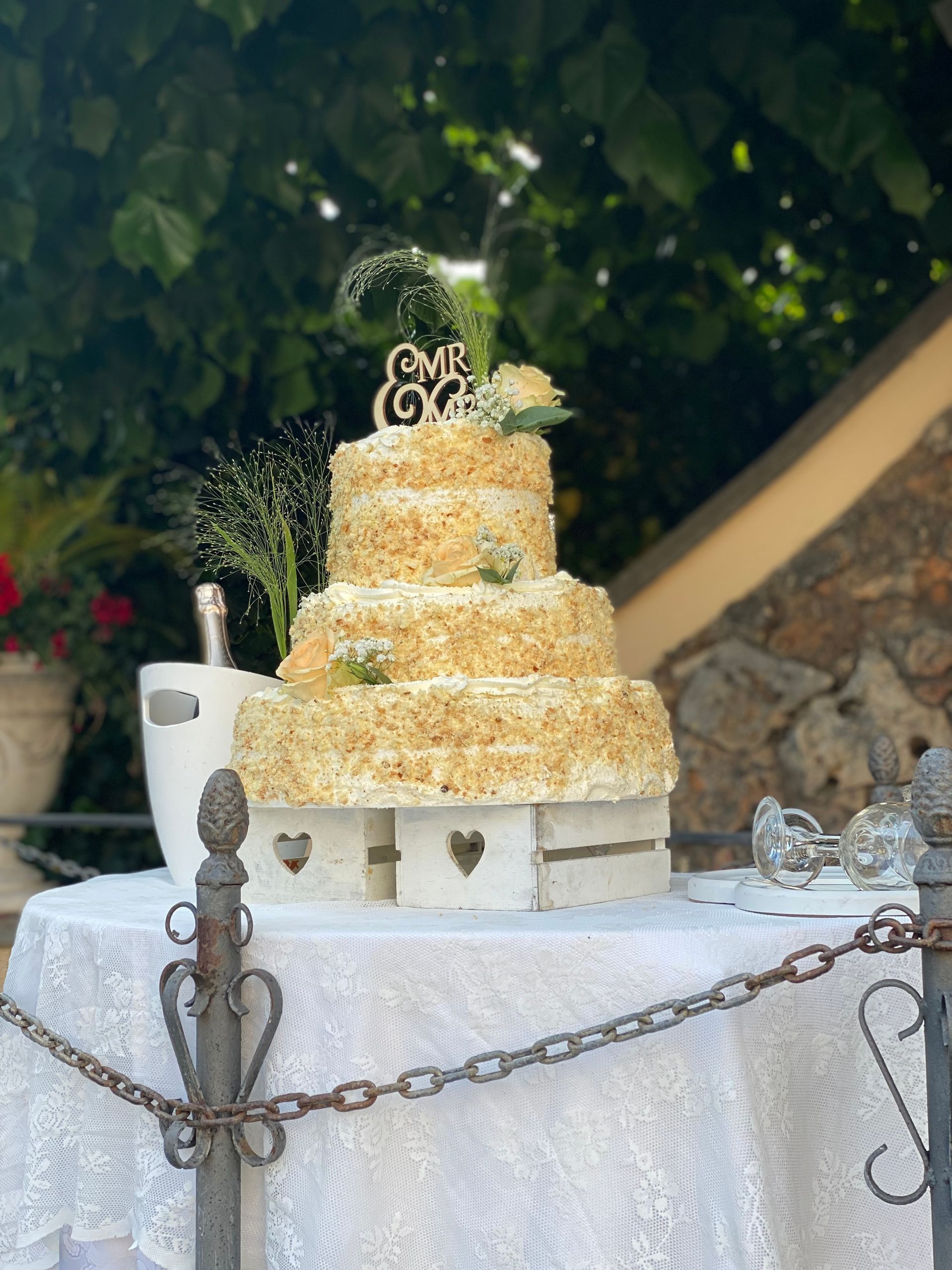 Torta nuziale a tre piani con glassa strutturata e decorazione su un tavolo con tovaglia bianca, all'aperto.