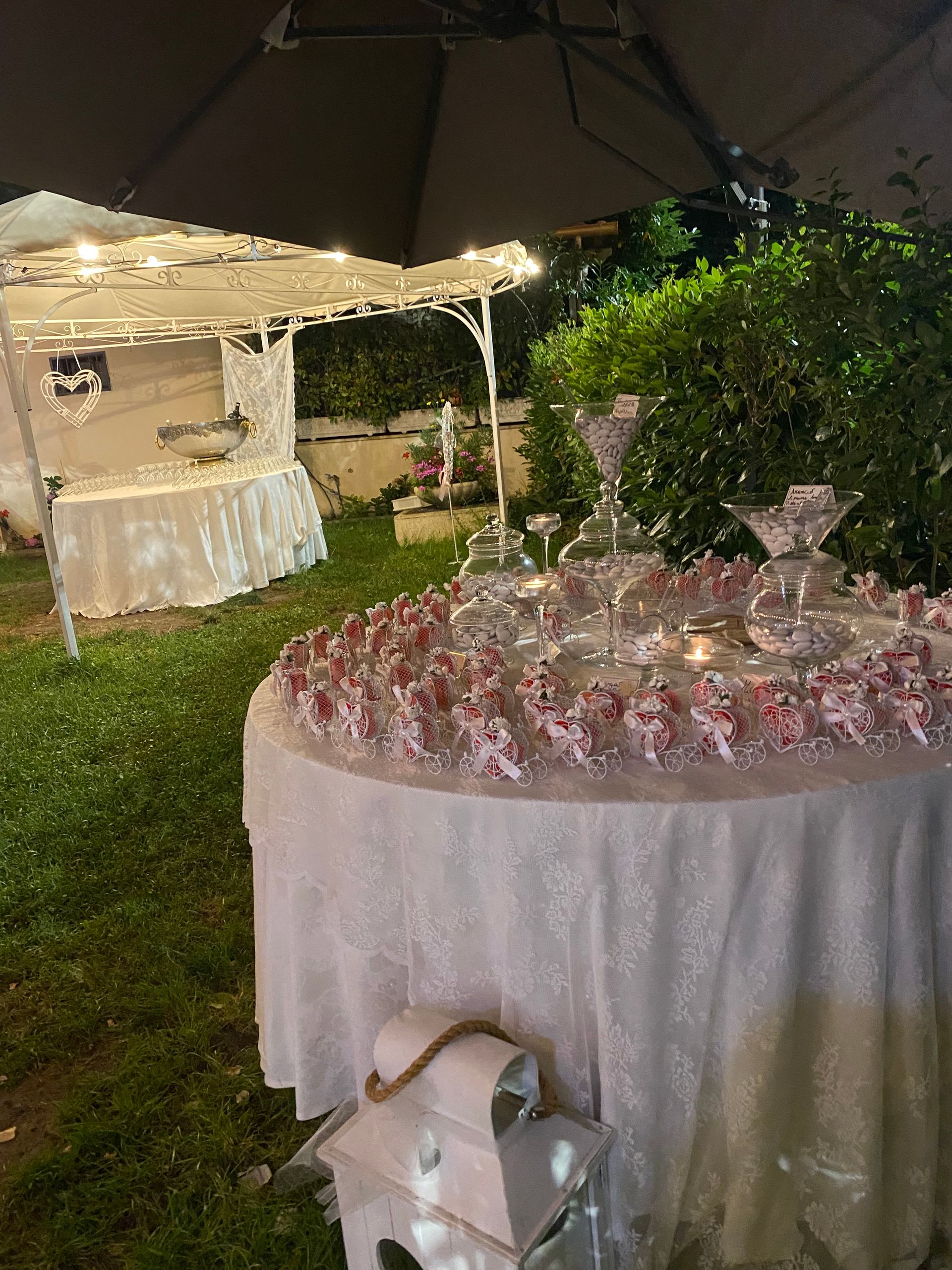 Ricevimento all'aperto: tavolo dei dolci con esposizione a più livelli in vetro di piccole prelibatezze, tovaglia bianca e gazebo sullo sfondo.