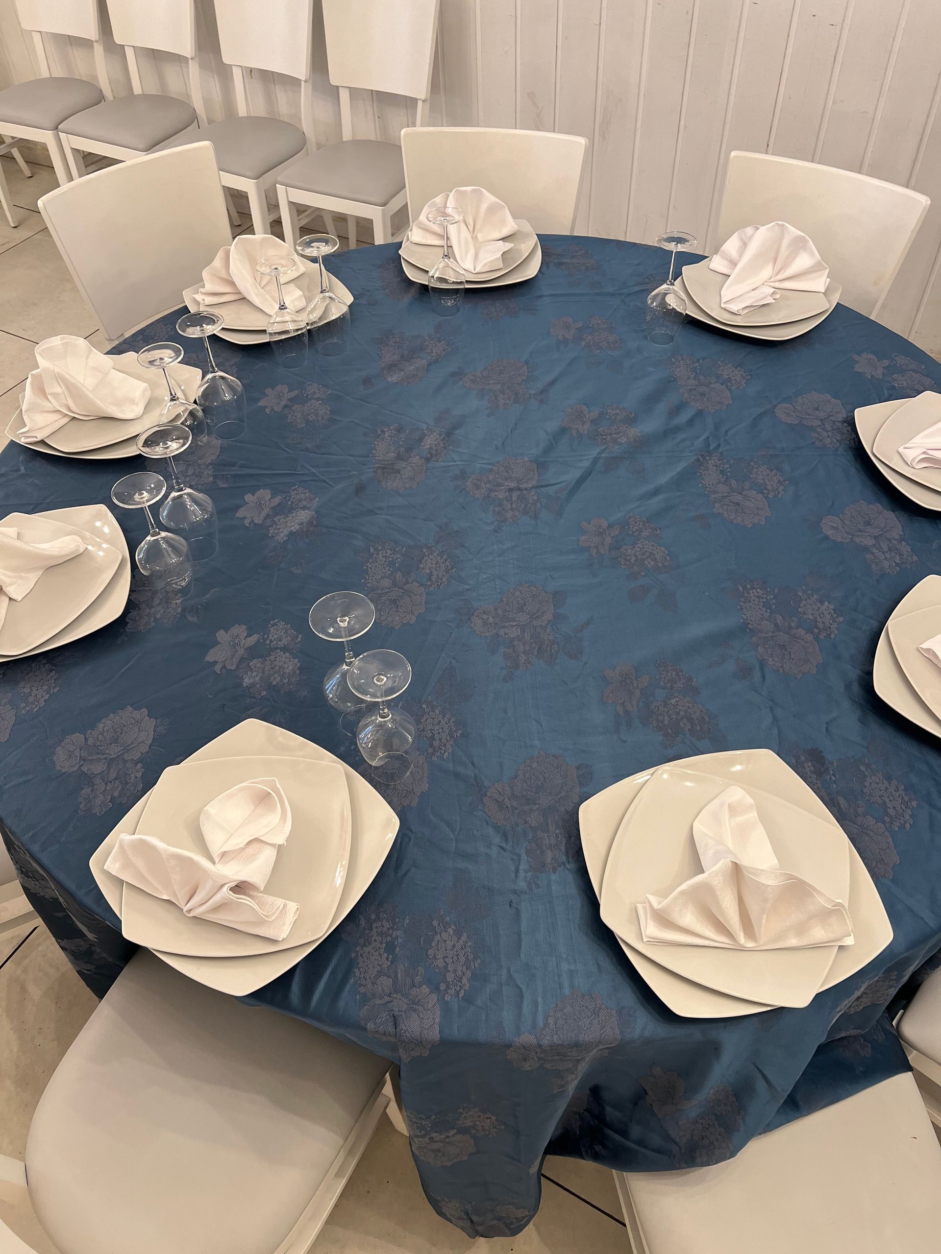 Tavolo rotondo apparecchiato per un pasto con piatti, tovaglioli e bicchieri. Tovaglia blu, sedie bianche, arredamento da interno.