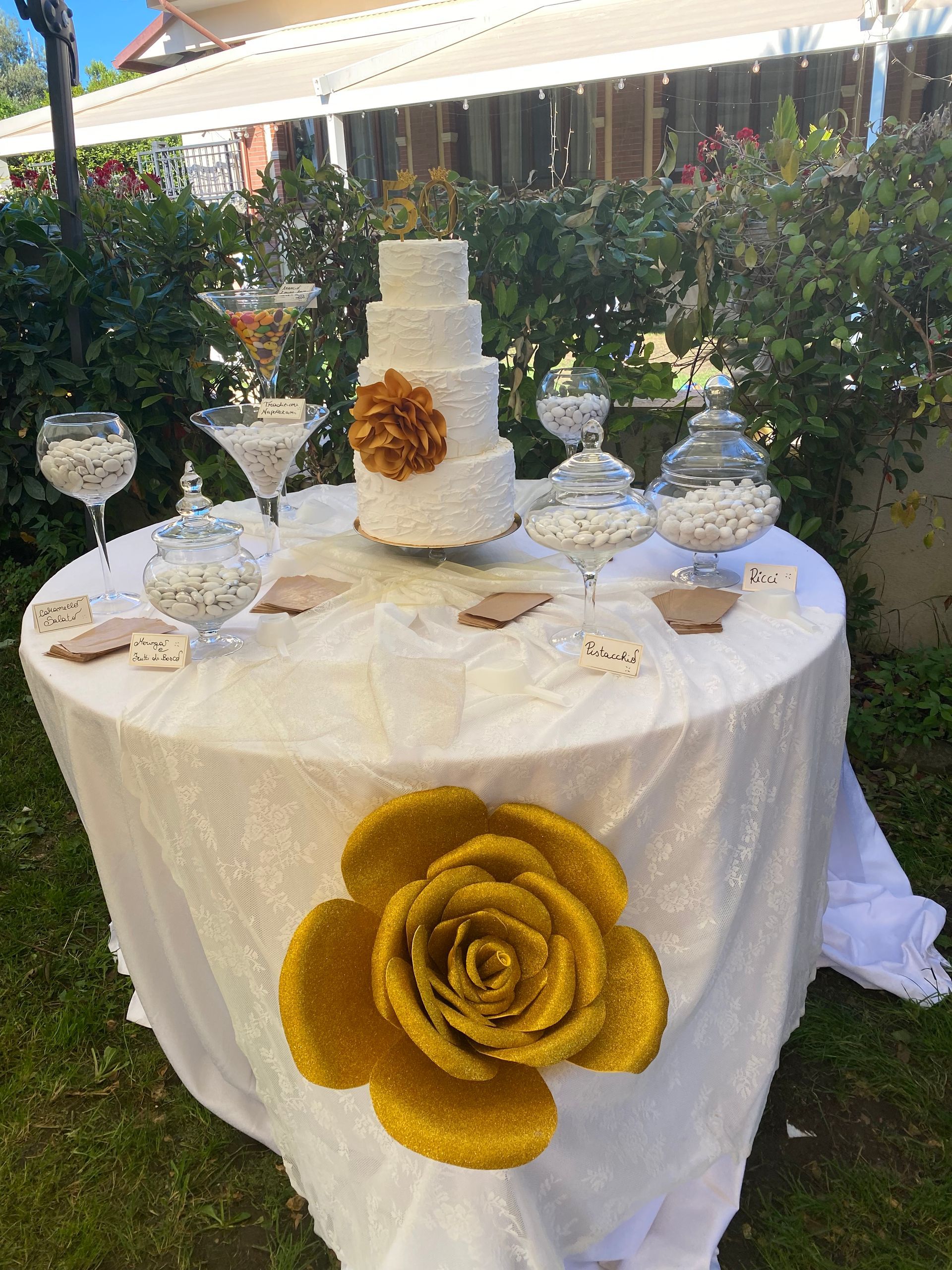 Tavolo bianco con torta nuziale, caramelle e decorazioni di rose dorate all'esterno.