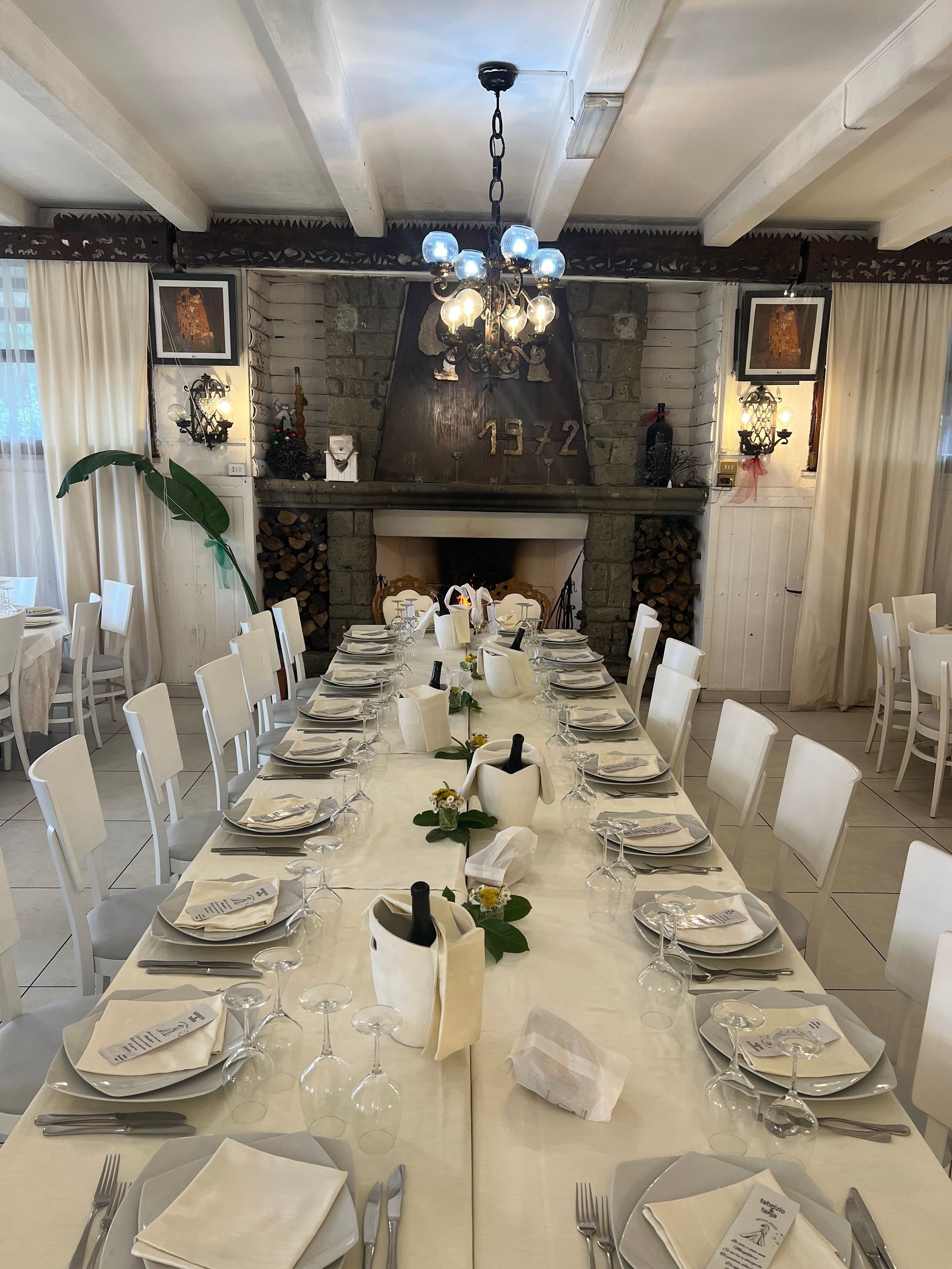 Lungo tavolo da pranzo apparecchiato per un pasto in un ristorante con decorazioni bianche e camino.