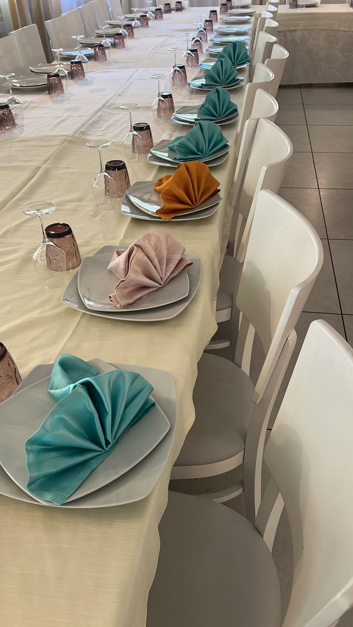 Lungo tavolo apparecchiato per un evento formale con sedie bianche, apparecchiato e tovaglioli colorati.
