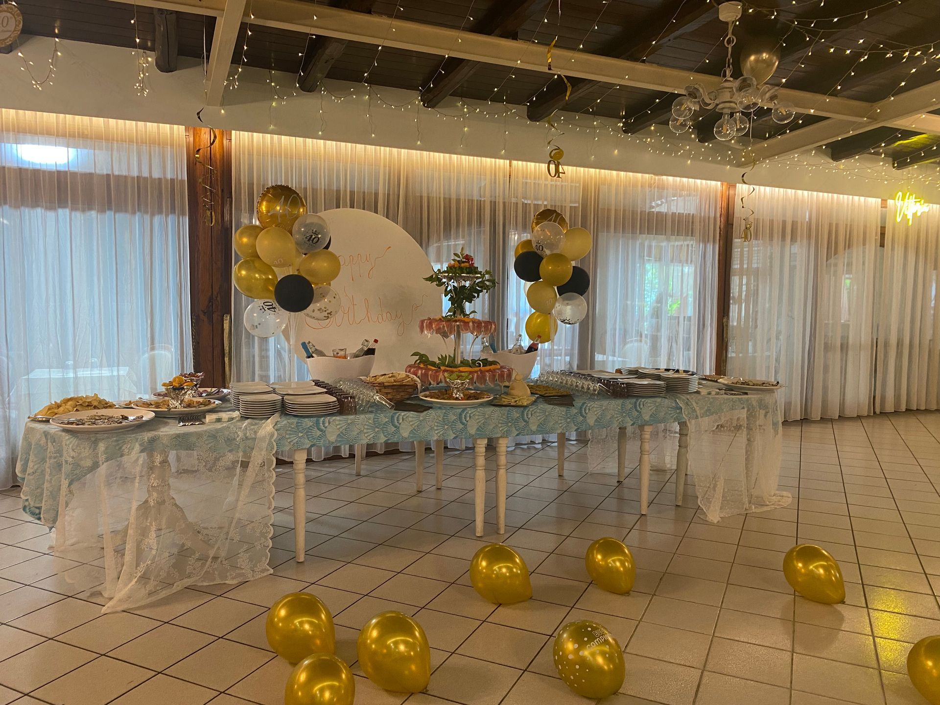 Tavolo da buffet apparecchiato con cibo, palloncini e decorazioni. Allestimento per festa al chiuso con tende bianche.