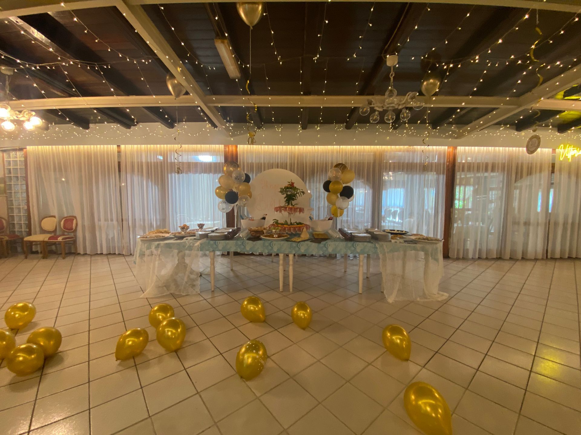 Tavolo apparecchiato con cibo e palloncini; palloncini dorati sul pavimento.