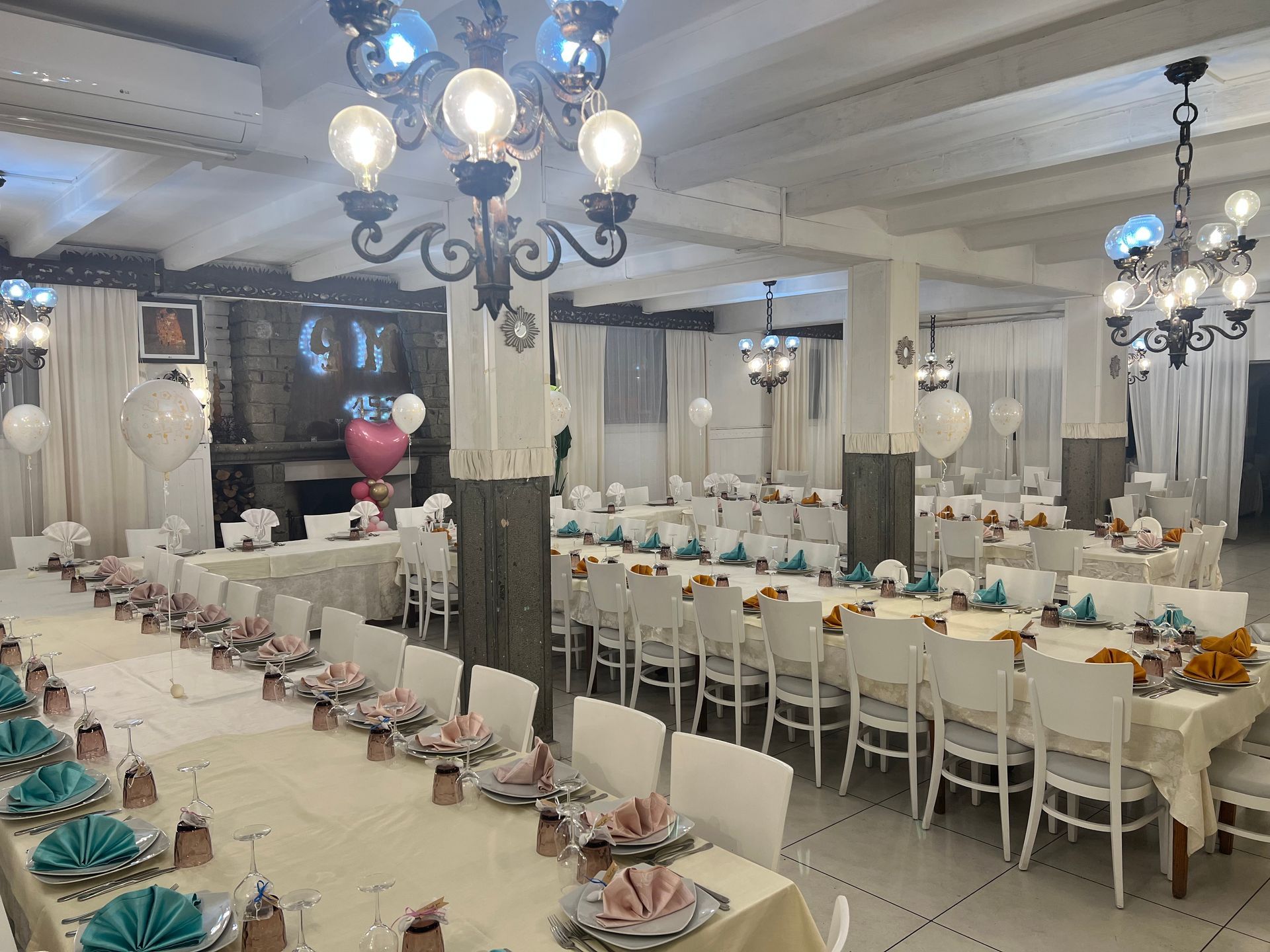 Sala per banchetti allestita con tavoli, sedie bianche e centrotavola floreali; lampadari in alto.