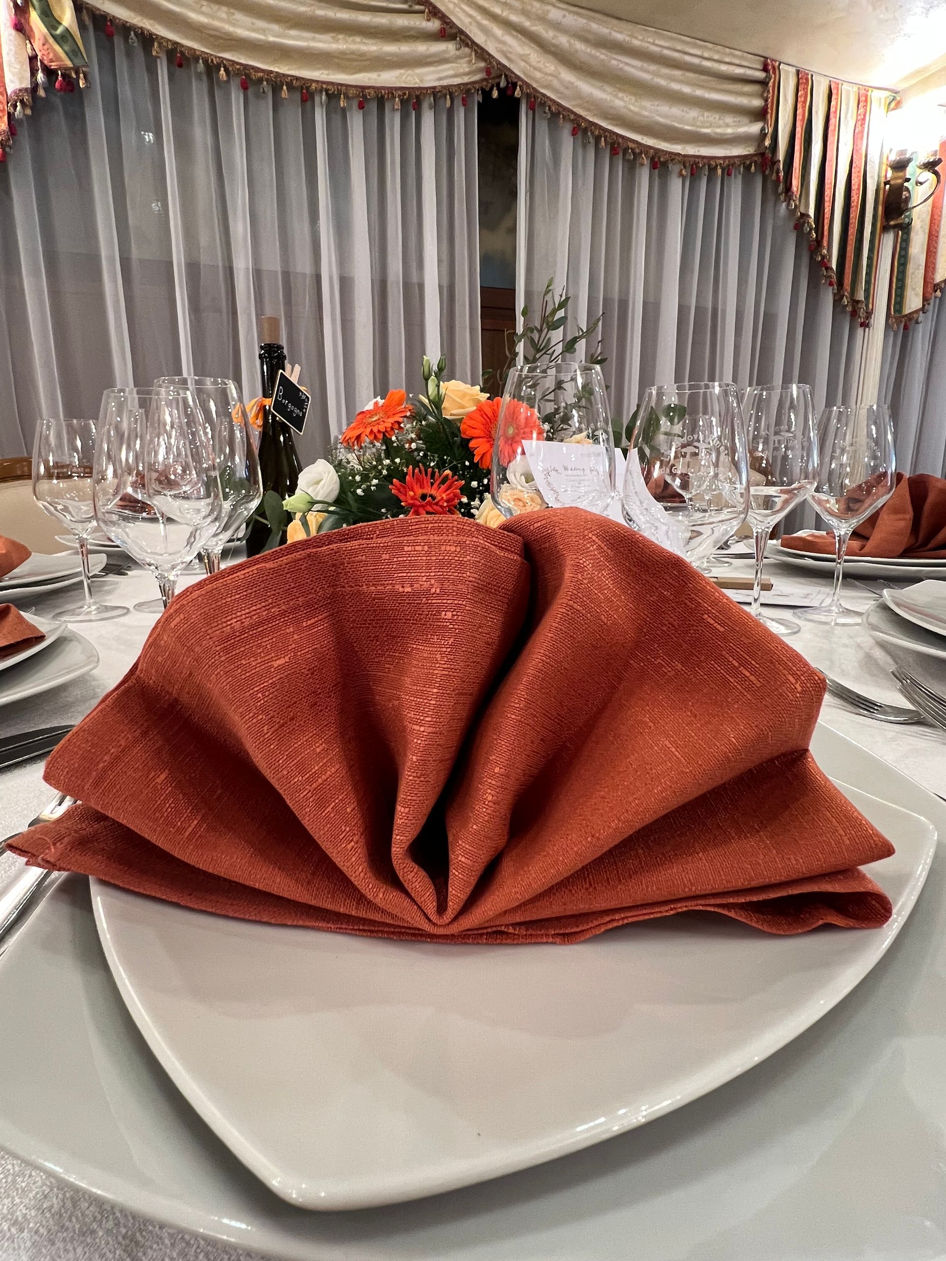 Apparecchiatura formale del tavolo con tovagliolo arancione piegato su un piatto bianco, centrotavola floreale e bicchieri da vino.