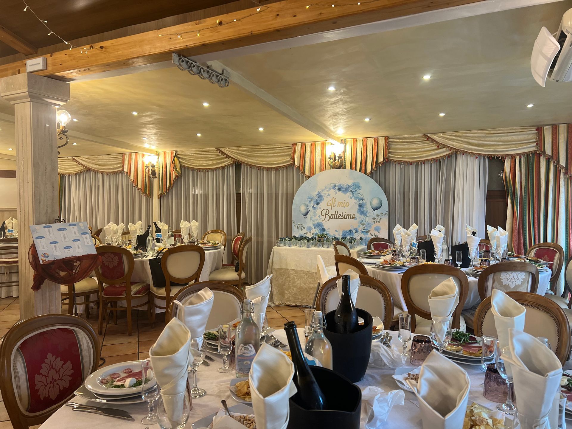Sala da pranzo formale allestita per un evento con tavoli decorati, tende e uno sfondo decorativo.