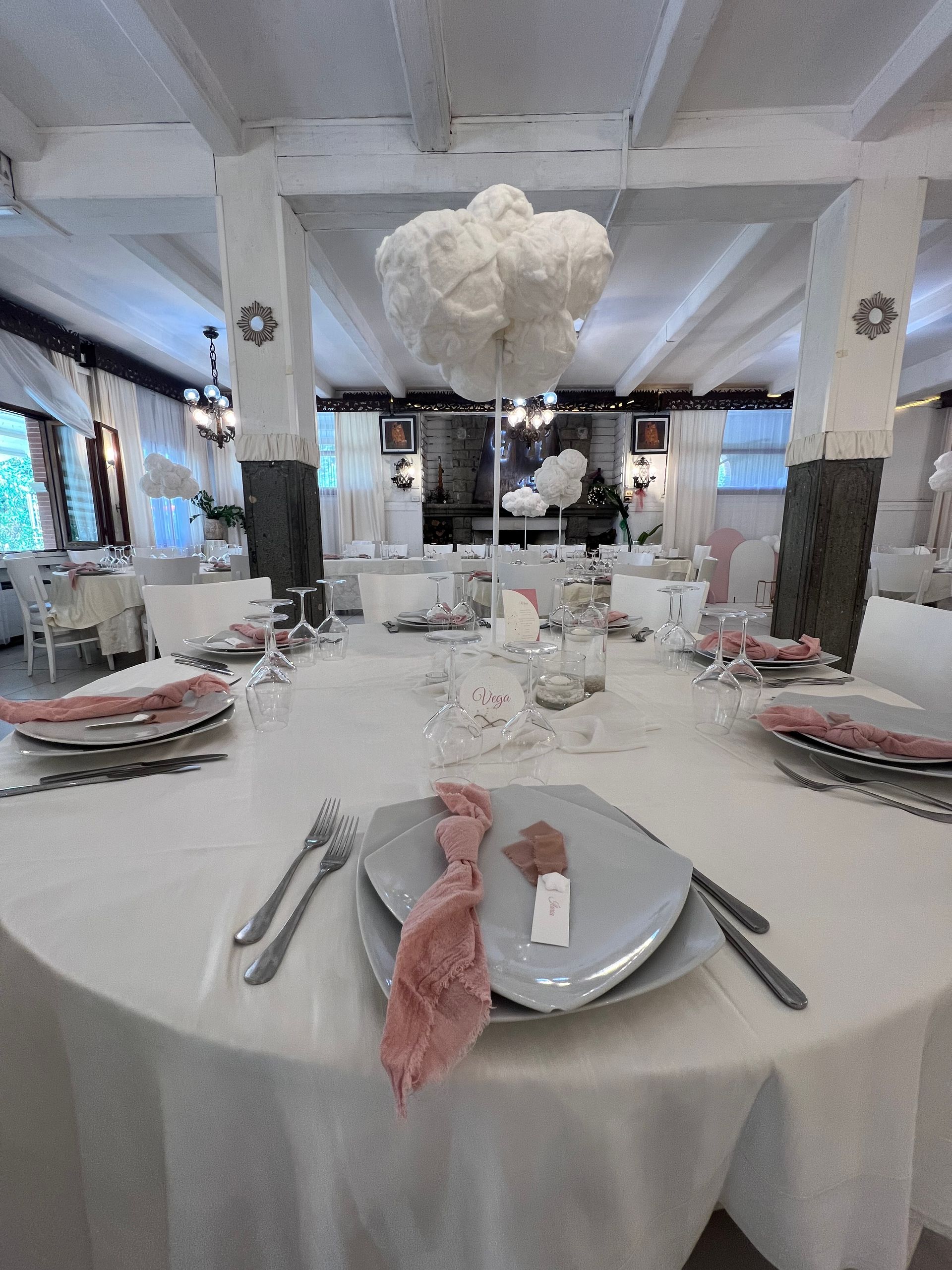 Tavolo rotondo apparecchiato per un evento formale, con tovaglie bianche e tovaglioli rosa, sotto una nuvola decorativa.
