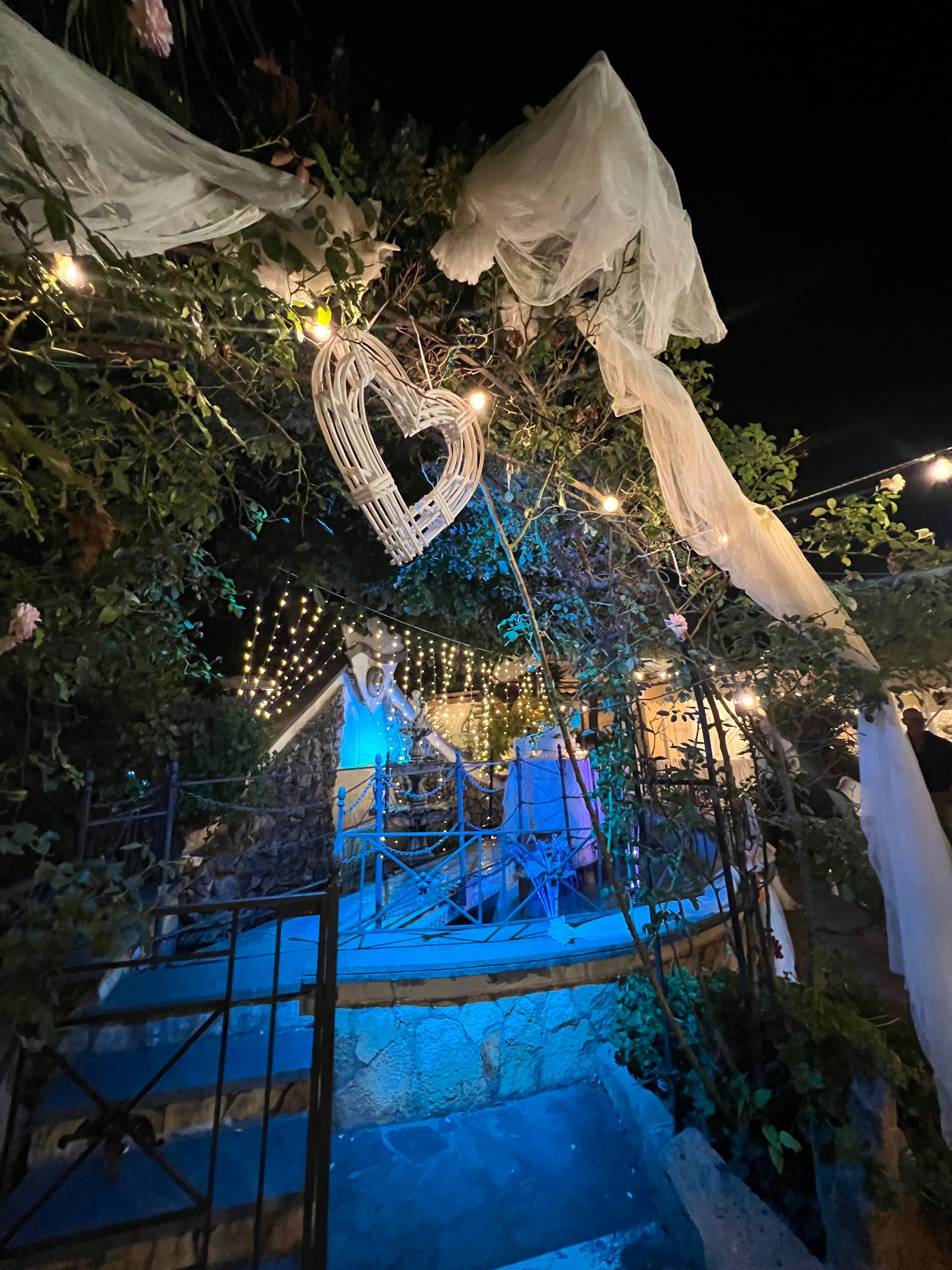 Arco da giardino notturno decorato con luci, tessuto e un ornamento a forma di cuore sopra un sentiero.