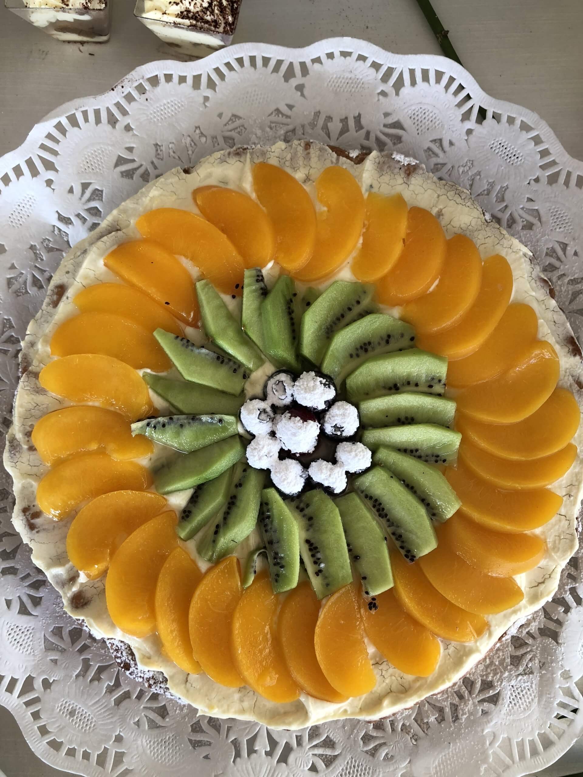 Crostata di frutta decorata con fette di pesca e kiwi disposte a forma di fiore, con glassa bianca.