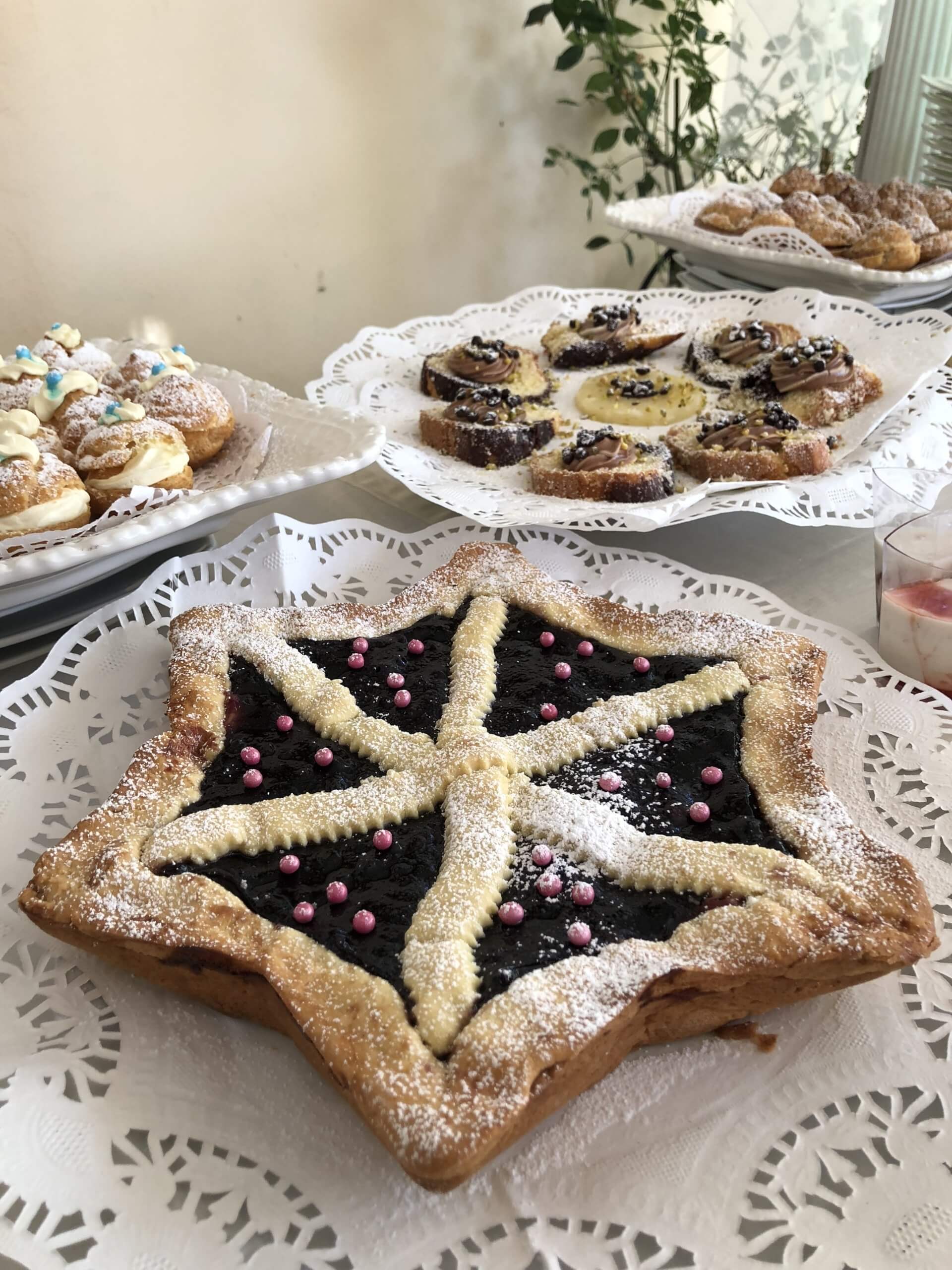 Pasta sfoglia a forma di stella con ripieno di frutti di bosco, spolverata di zucchero a velo, su un centrino di pizzo, con altri dolci sullo sfondo.