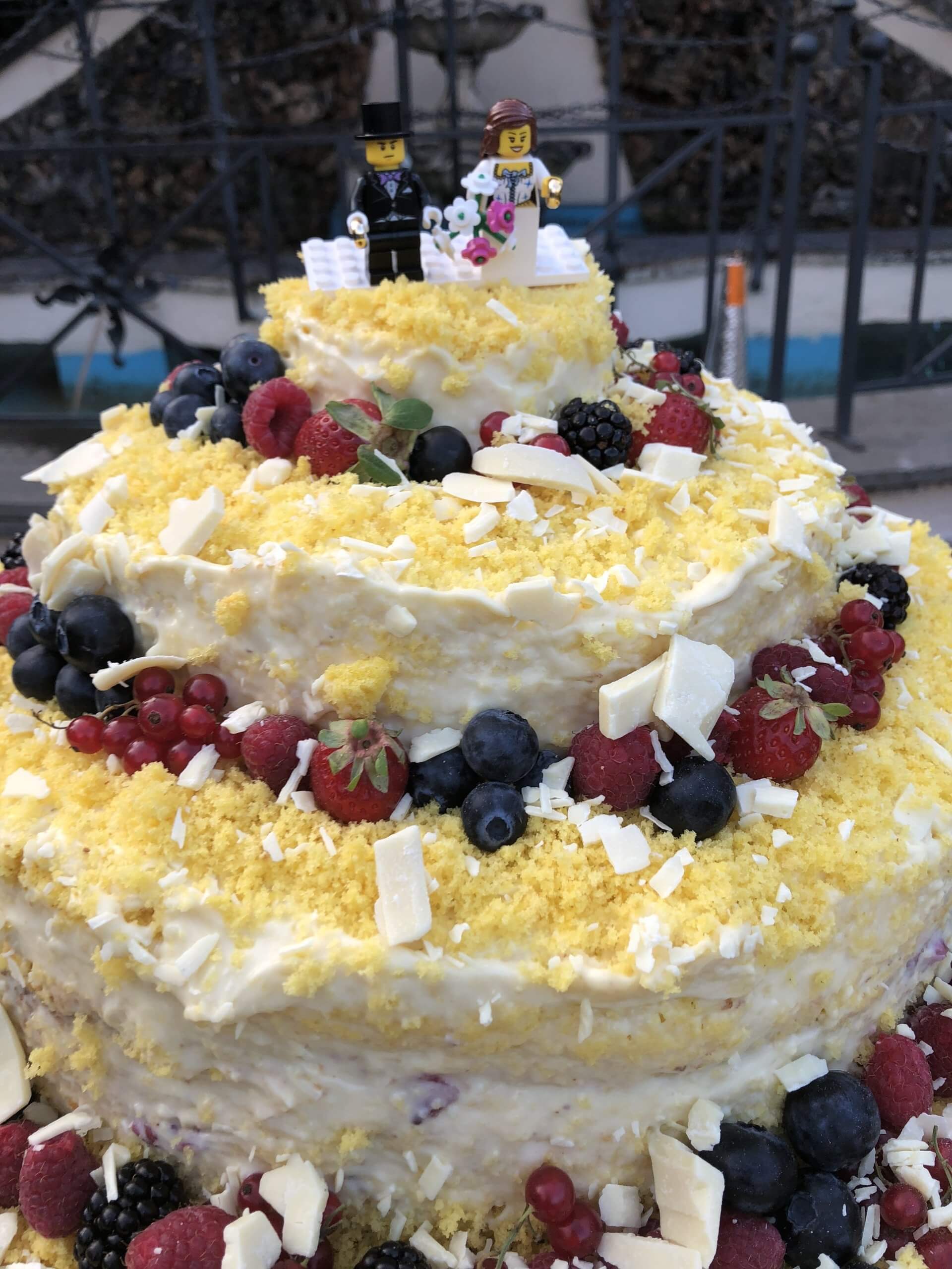 Torta nuziale a tre piani con decorazioni a forma di sposi Lego, guarnita con frutti di bosco e scaglie di cioccolato bianco.