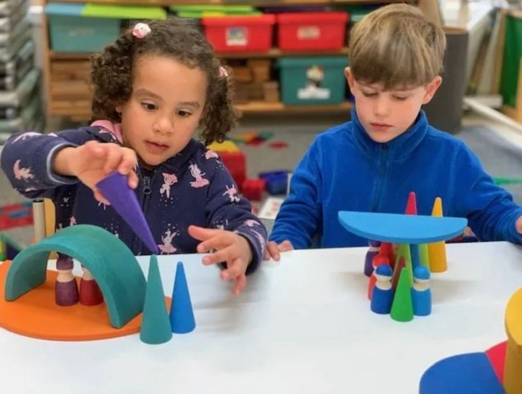Child with hat smiles from inside a playhouse with colorful cups on a ledge.
