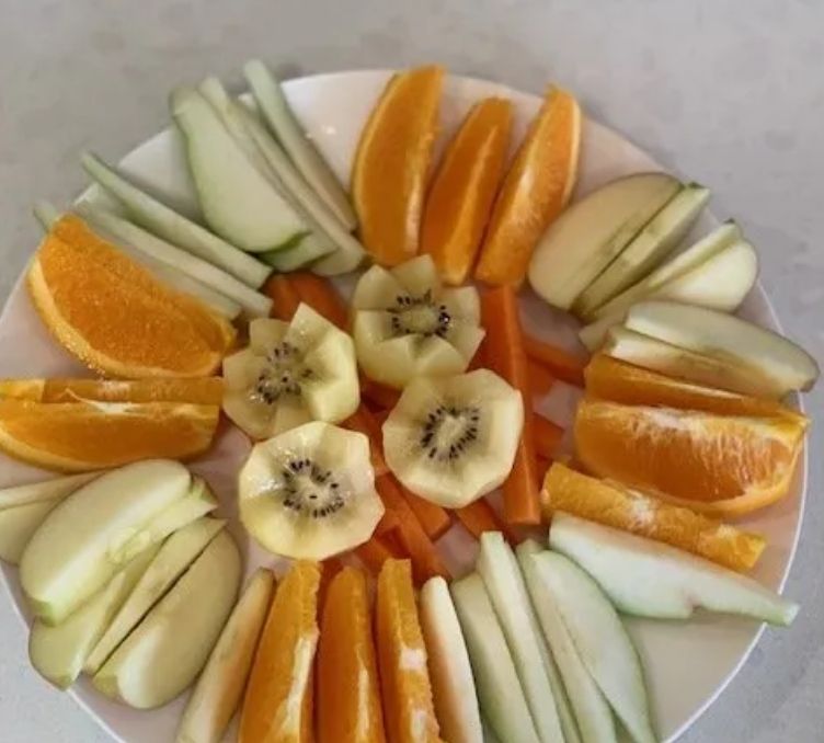 A plate of sliced fruits and vegetables: oranges, kiwi, apples, carrots, and cucumbers.