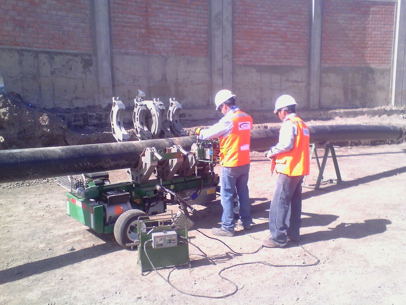 Dos trabajadores con chalecos y cascos de seguridad manejan maquinaria pesada para cortar tuberías al aire libre.