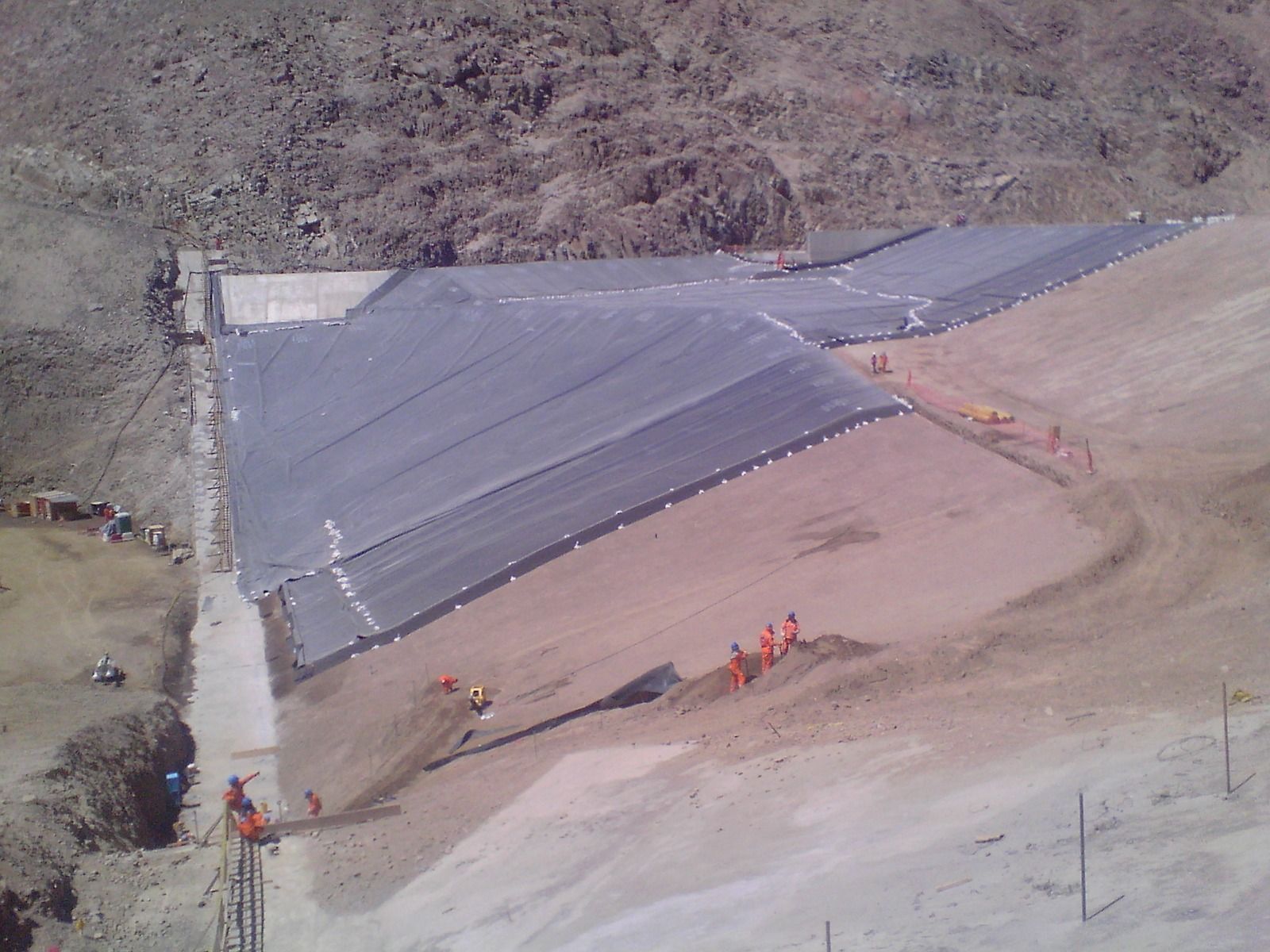 Obra en construcción con una gran lona gris que cubre un terraplén inclinado y trabajadores con chalecos naranjas cerca.