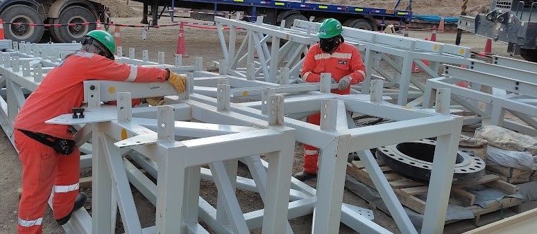 Trabajadores con monos naranjas y cascos verdes ensamblan una estructura metálica blanca al aire libre.