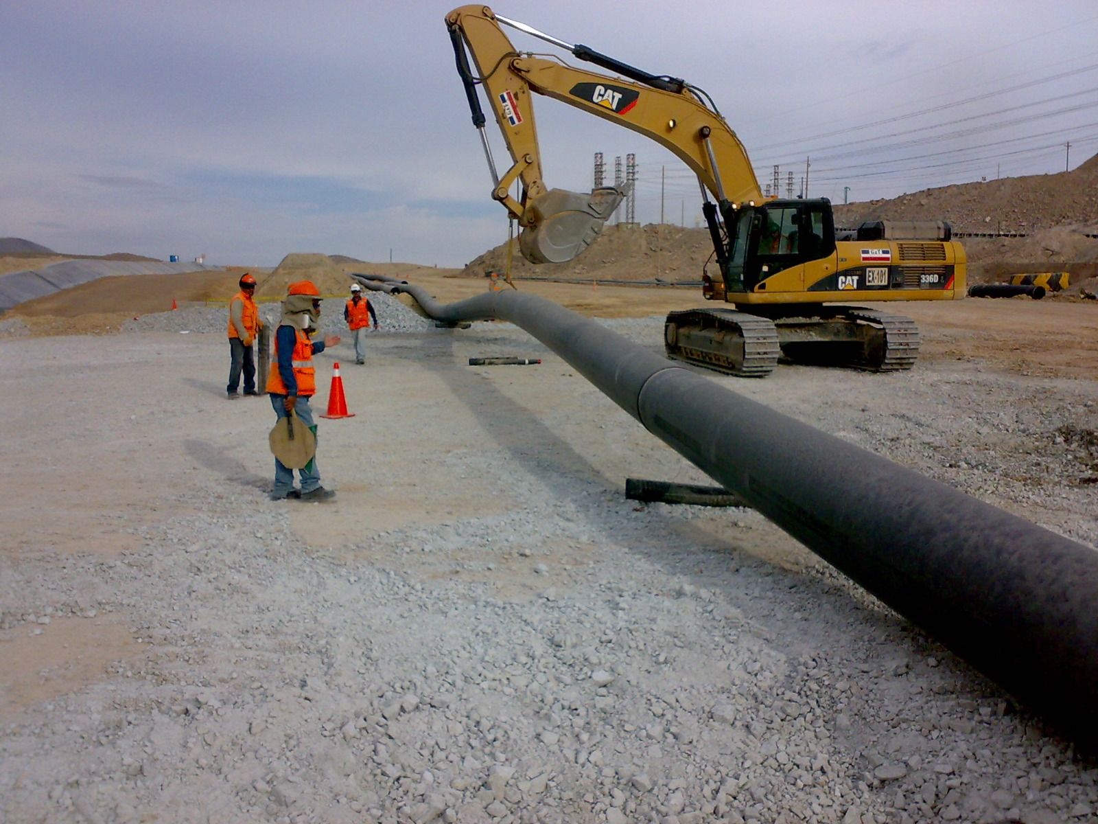 Obreros de la construcción y una excavadora colocan una gran tubería negra en un terreno de grava.