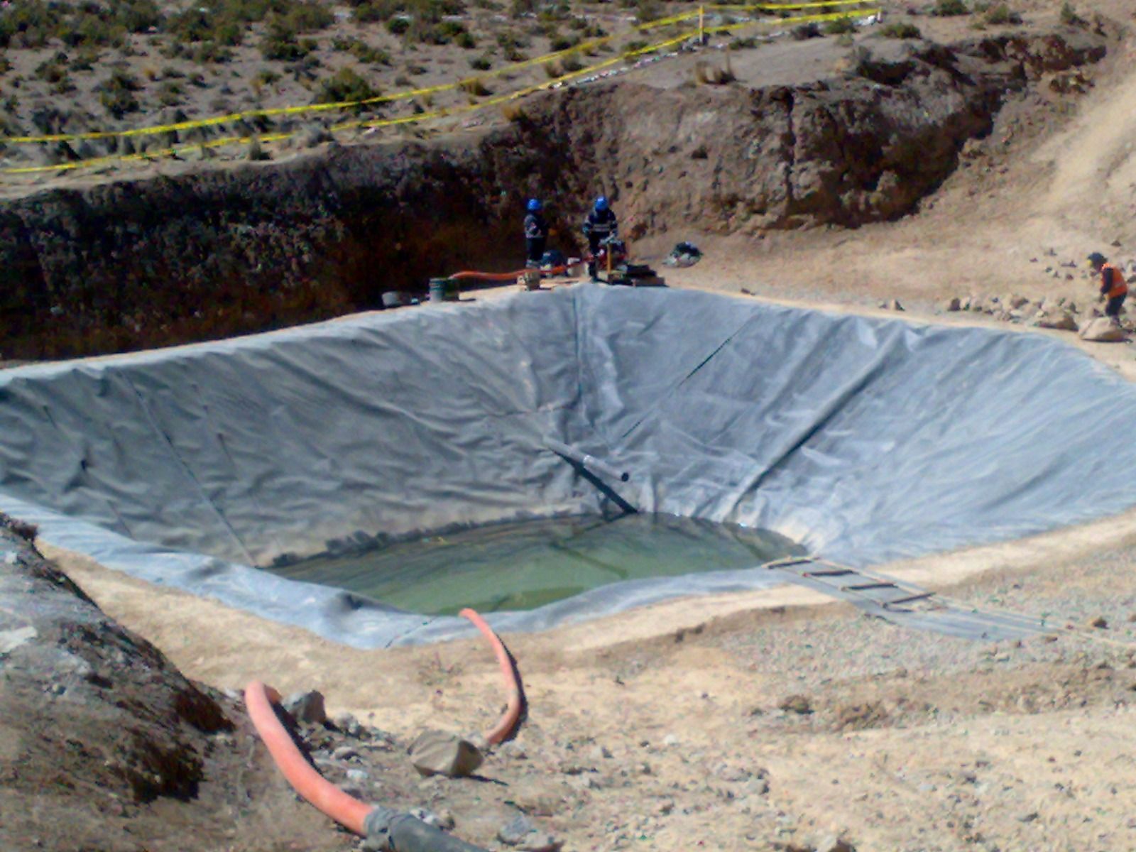 Fosa de excavación revestida con una pequeña poza de agua y trabajadores en el borde en un sitio rocoso desértico.