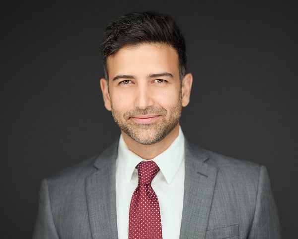 Man in gray suit, white shirt, and red polka dot tie smiles at the camera, against a dark gray background.