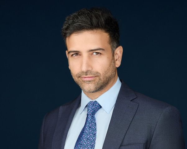 Man in a suit, blue tie, looking at the camera against a dark blue background.