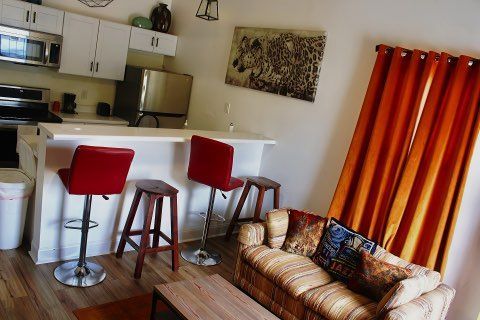 A small kitchen with a breakfast bar and red stools opens to a living area with a couch and orange curtains.