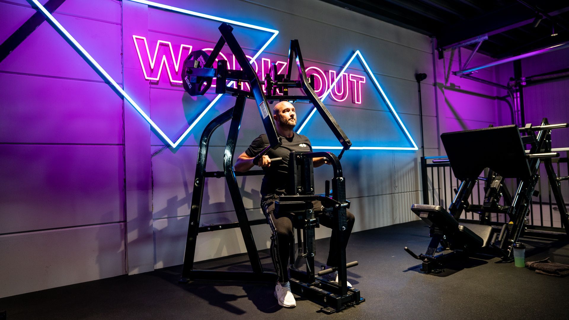 Een man zit op een apparaat in een sportschool onder een neonreclame.