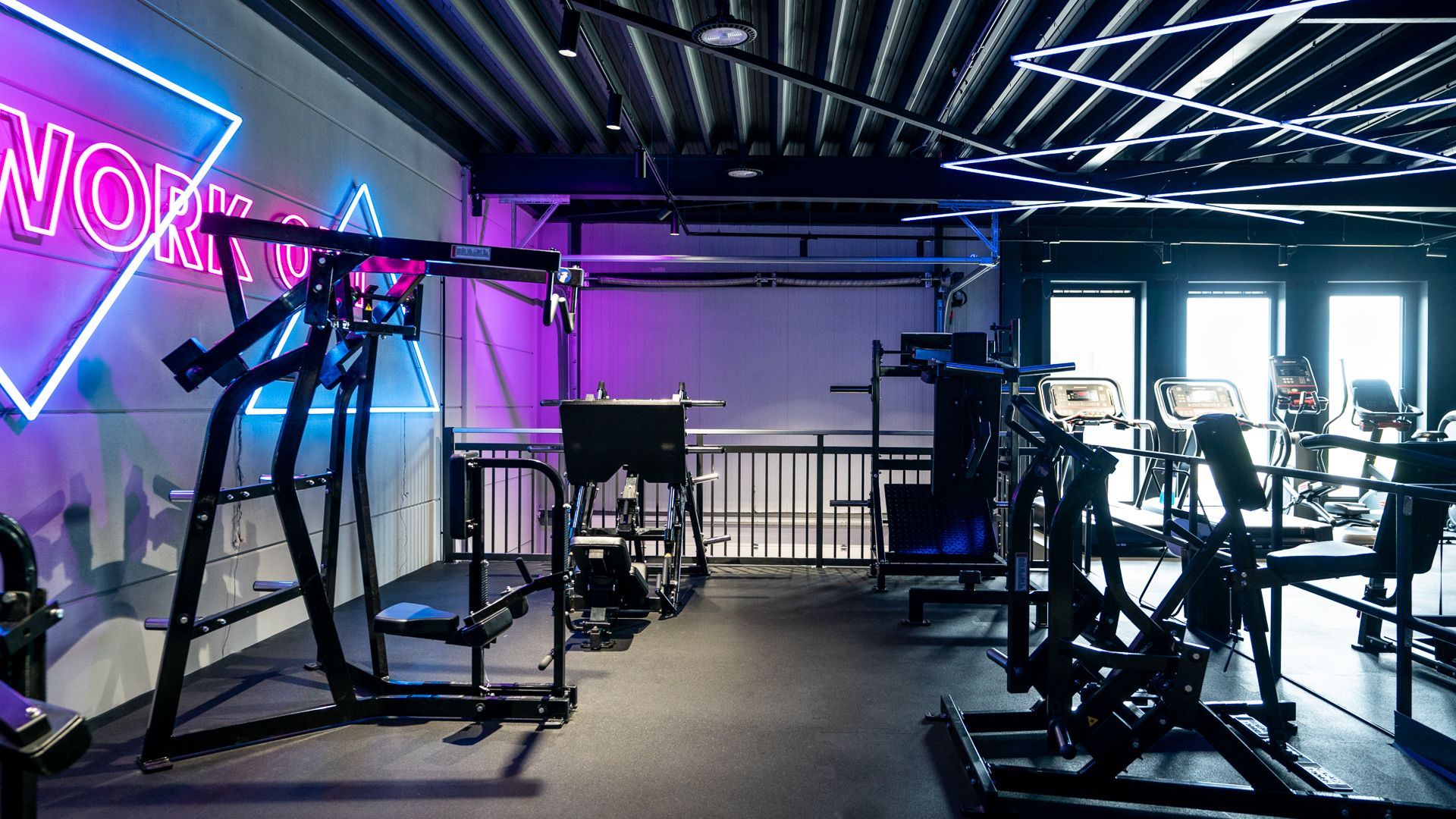Een sportschool met veel fitnessapparatuur en een neonreclame aan de muur.