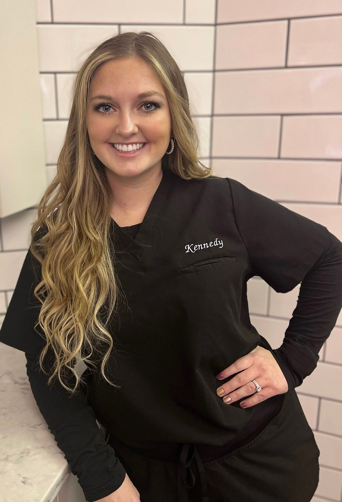 Woman in black scrubs, smiling, leaning on a counter, against a white tile wall.