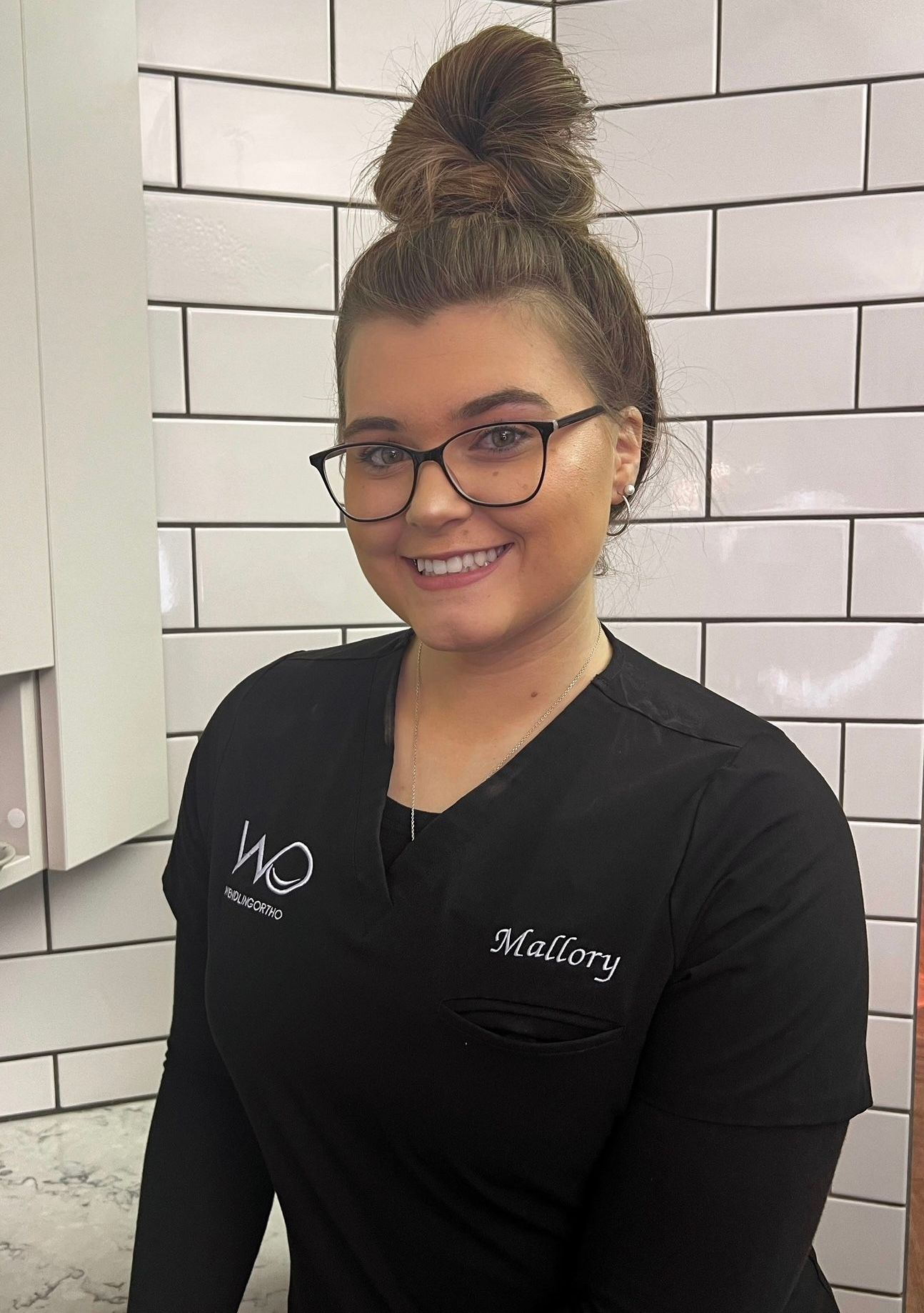 A woman wearing glasses and a black scrub top is smiling in front of a white brick wall.