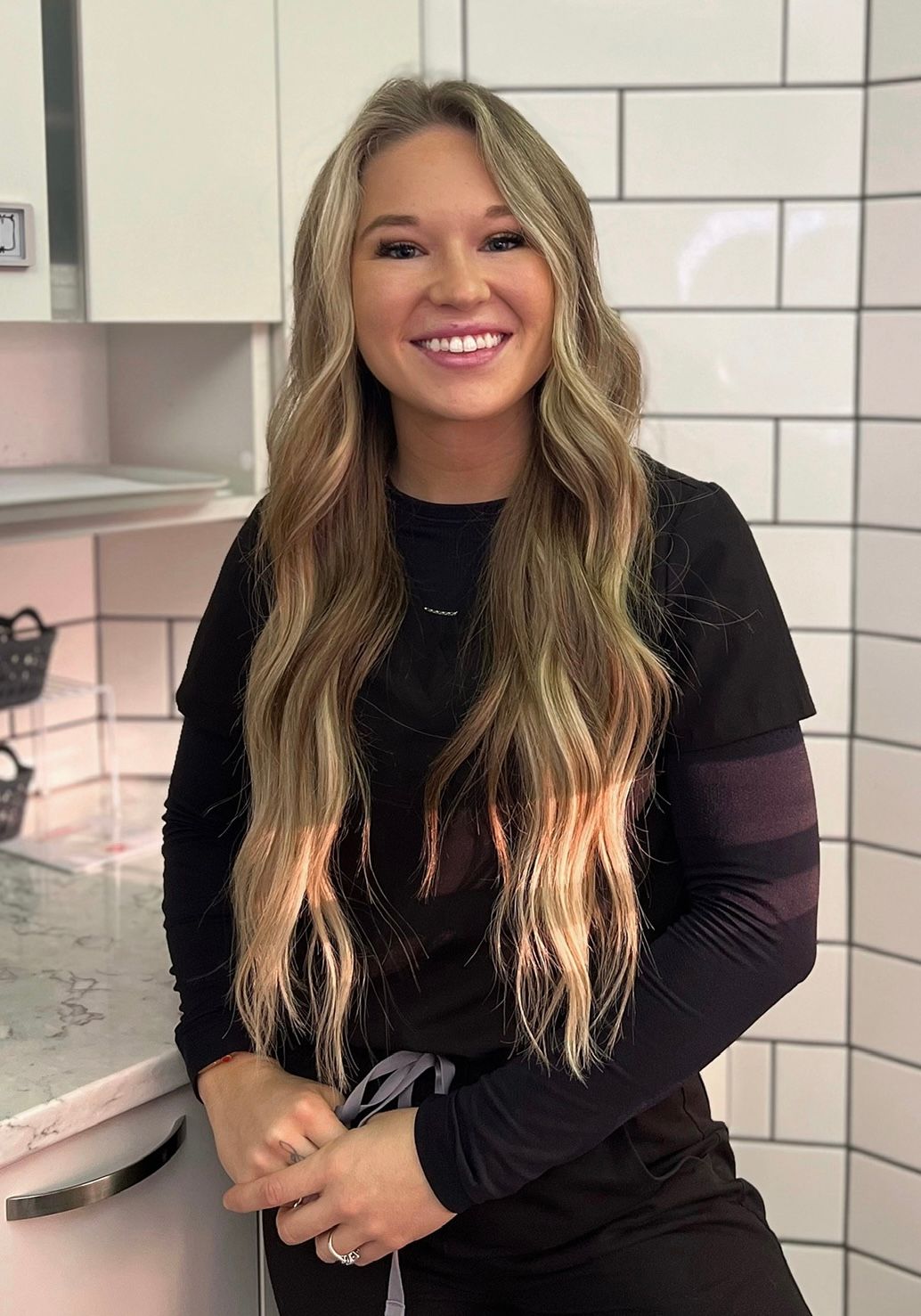 A woman with long blonde hair is smiling for the camera in a kitchen.