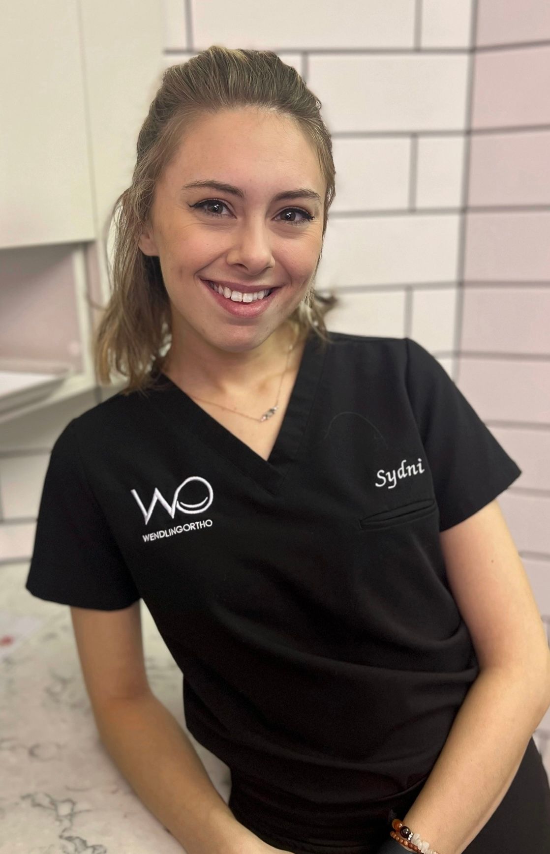 A woman in a black scrub top is smiling and sitting on a counter.