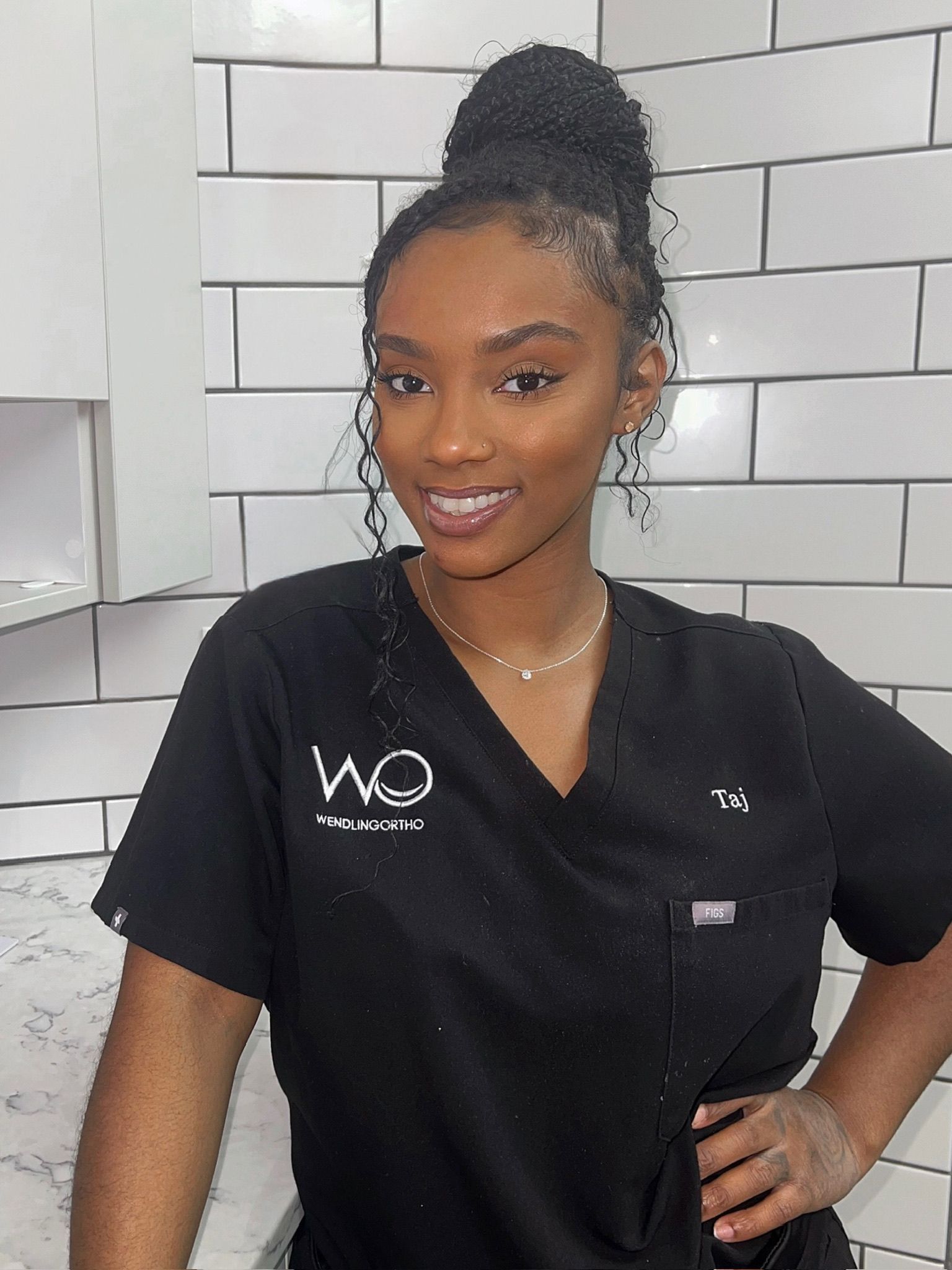 A woman wearing a black scrub top is standing in front of a white brick wall.