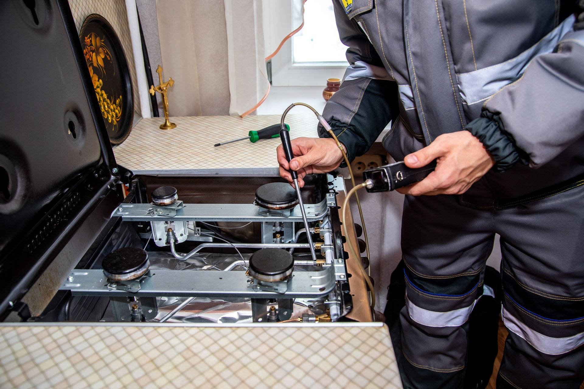 A person in work clothes repairing a stovetop, holding tools over the burners.