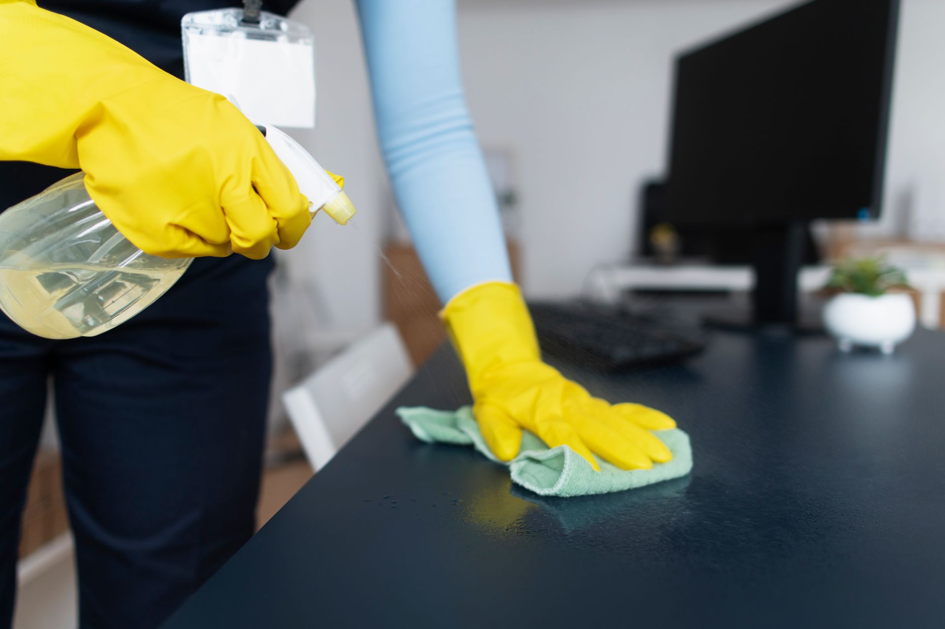 A person wearing yellow gloves is cleaning a desk with a cloth and spray bottle.