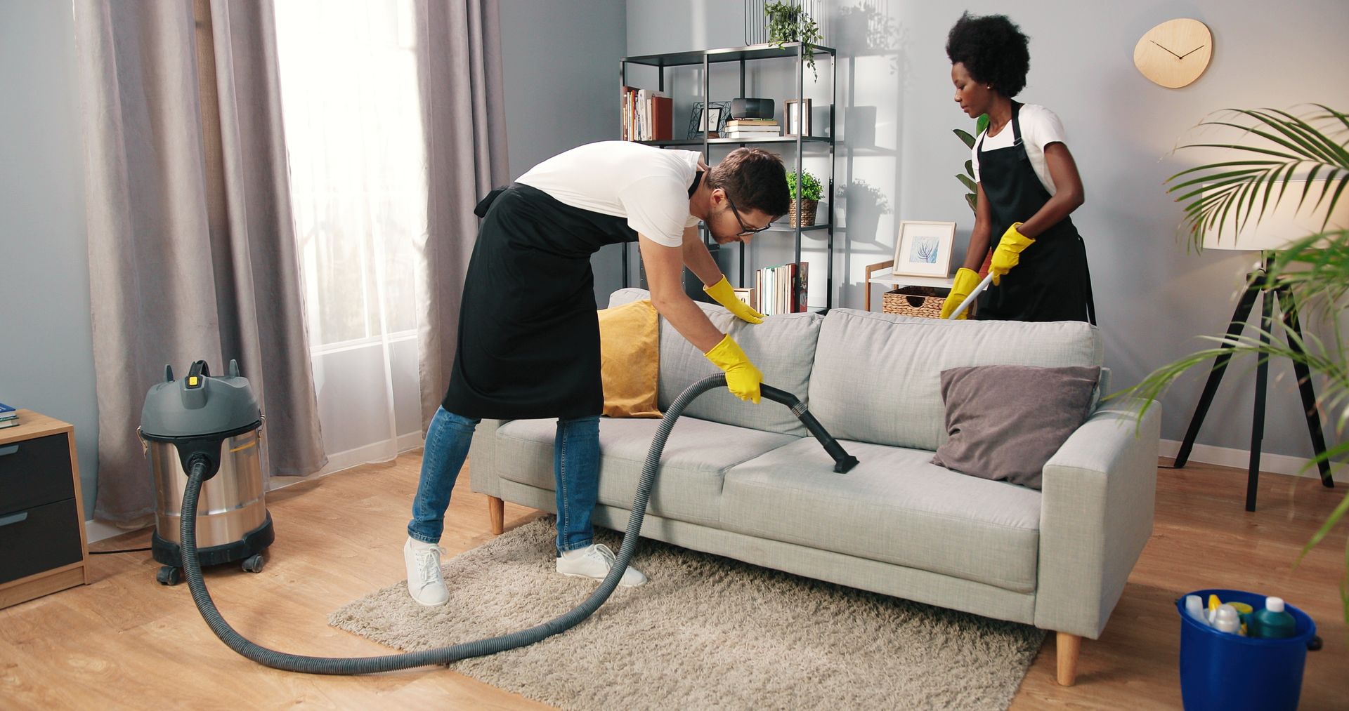 A man and a woman are cleaning a couch with a vacuum cleaner.