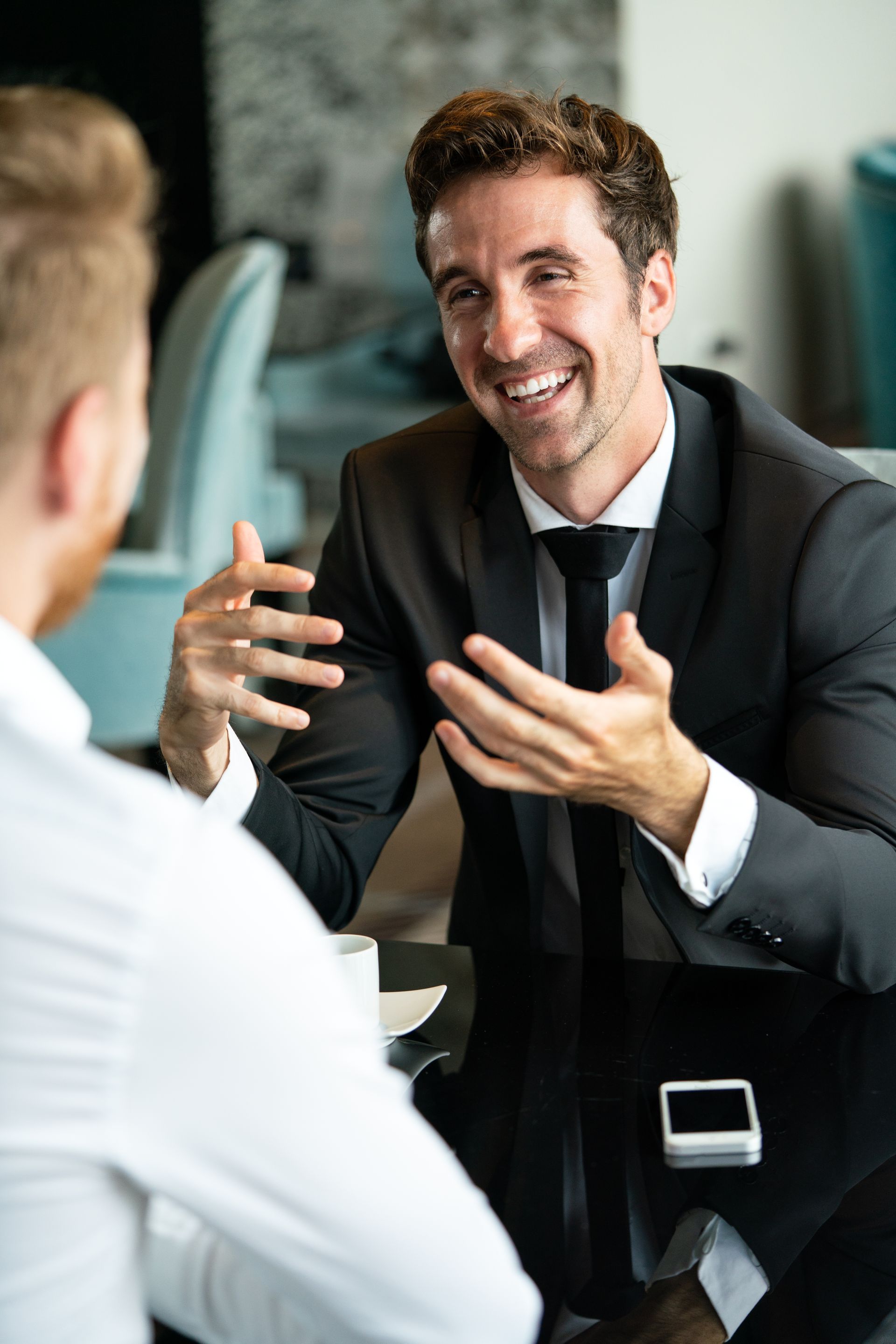 A man in a suit and tie is sitting at a table talking to another man.