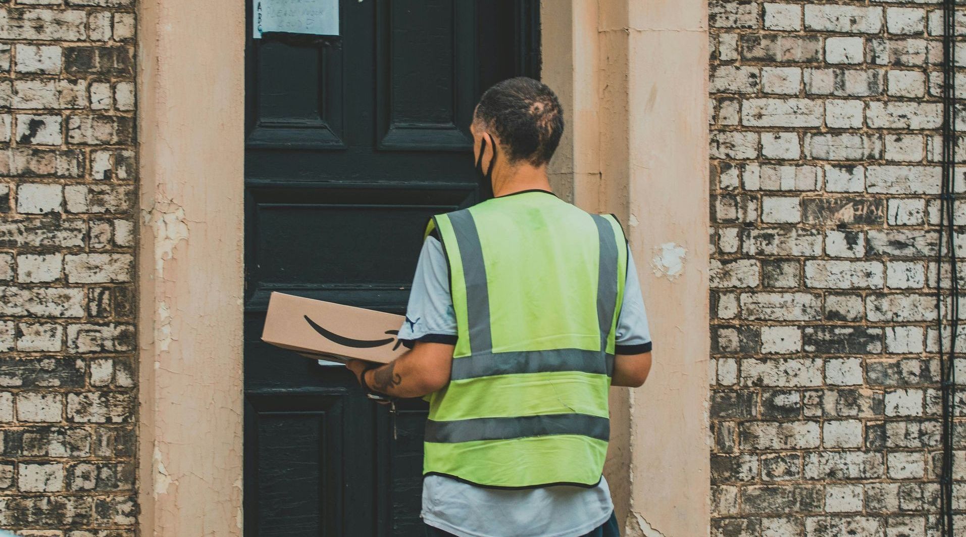 A man in a yellow vest is holding a cardboard box in front of a door.