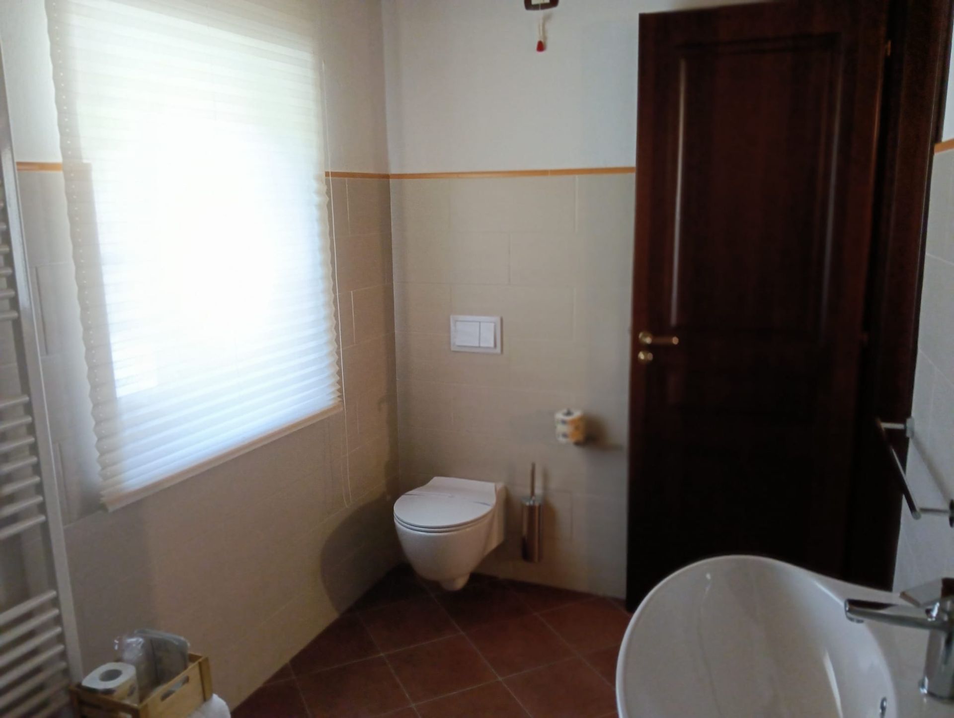 Bagno con WC, finestra e porta marrone scuro. Pareti beige con pavimento in piastrelle marroni.