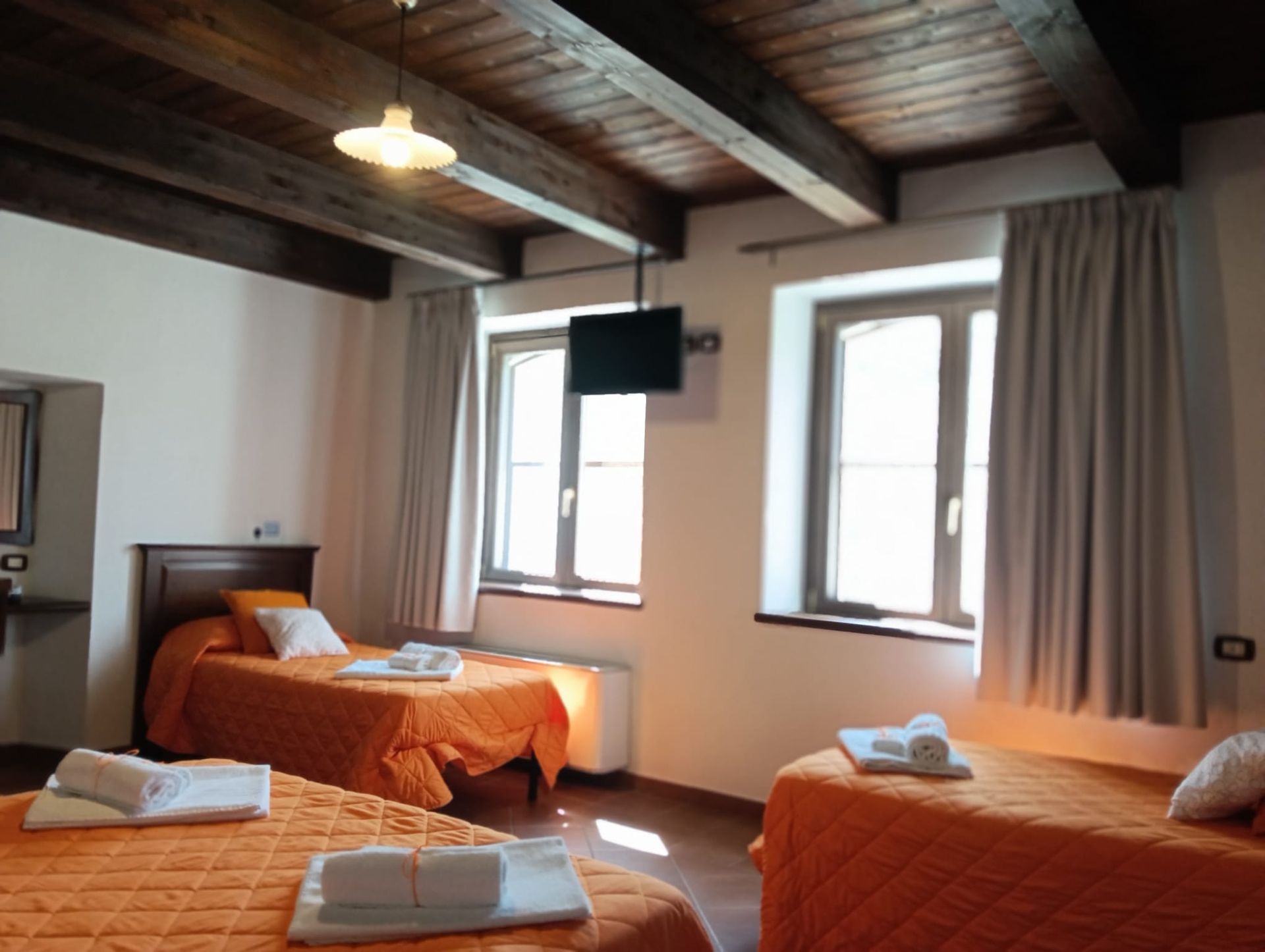 Camera da letto con tre letti, biancheria arancione, travi in legno e finestre con tende grigie.