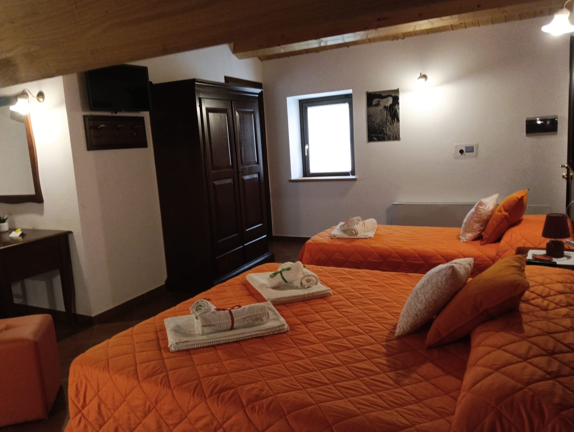 Camera da letto con tre letti arancioni, armadio in legno scuro, piccola finestra, pareti bianche e soffitto in legno.