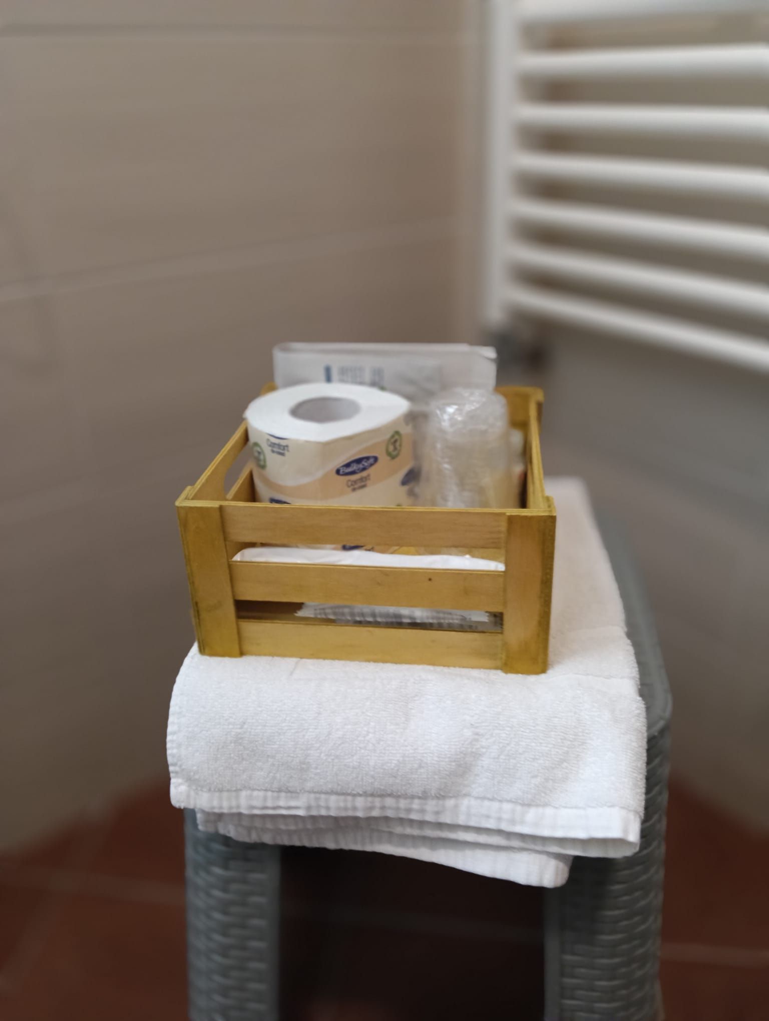 Cassa di legno con carta igienica, bicchieri e sapone su un asciugamano bianco piegato, appoggiato su uno sgabello grigio.