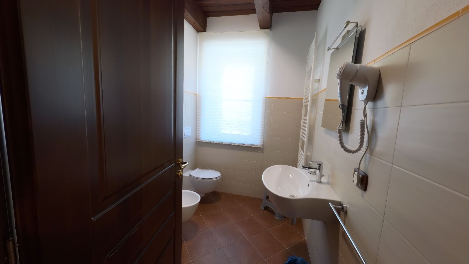 Interno del bagno con lavandino, WC e finestra. Porta marrone sulla sinistra e asciugacapelli a parete.
