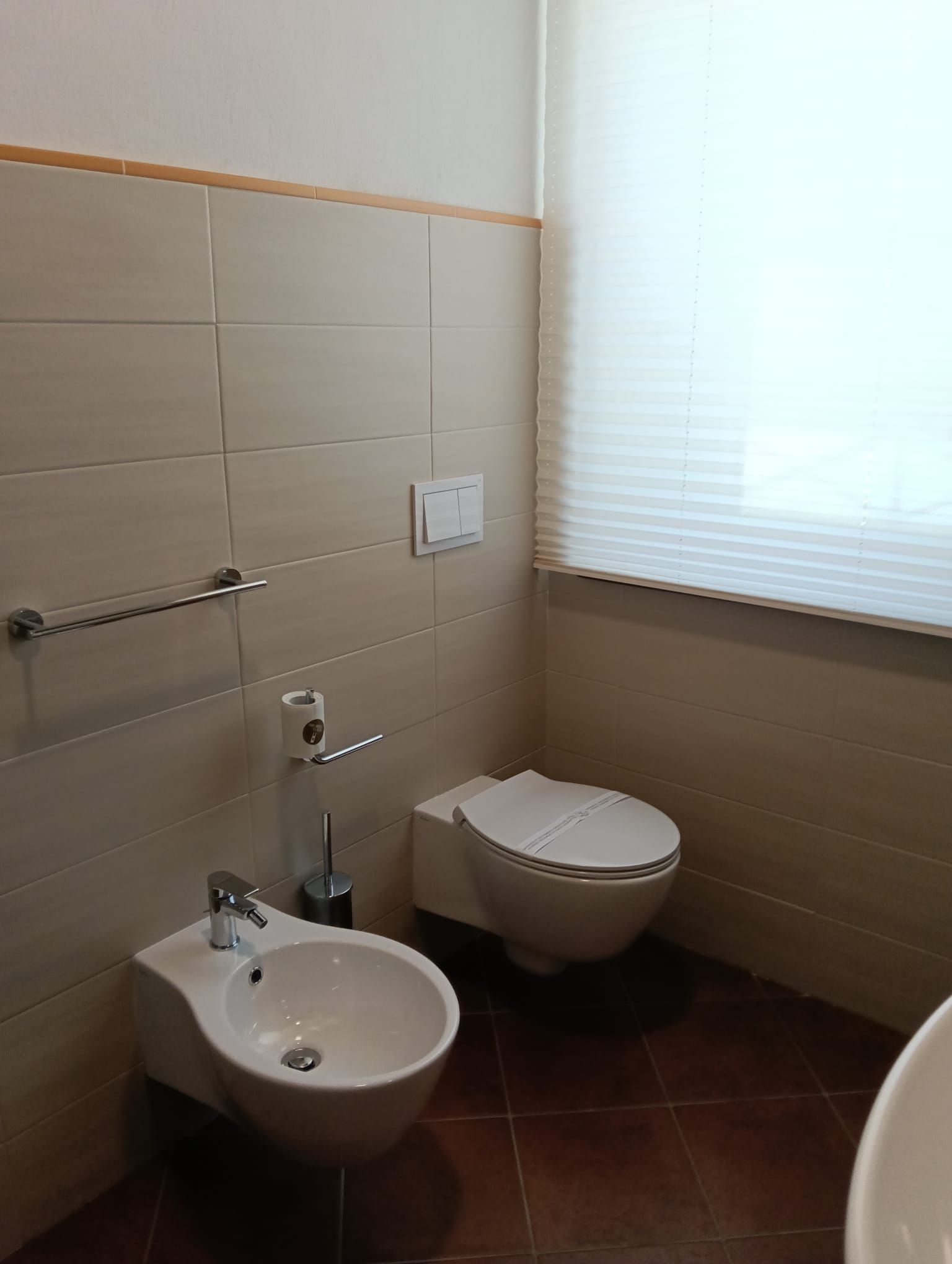 Bagno con bidet, WC e piastrelle bianchi. Pavimento marrone e finestra con persiane.