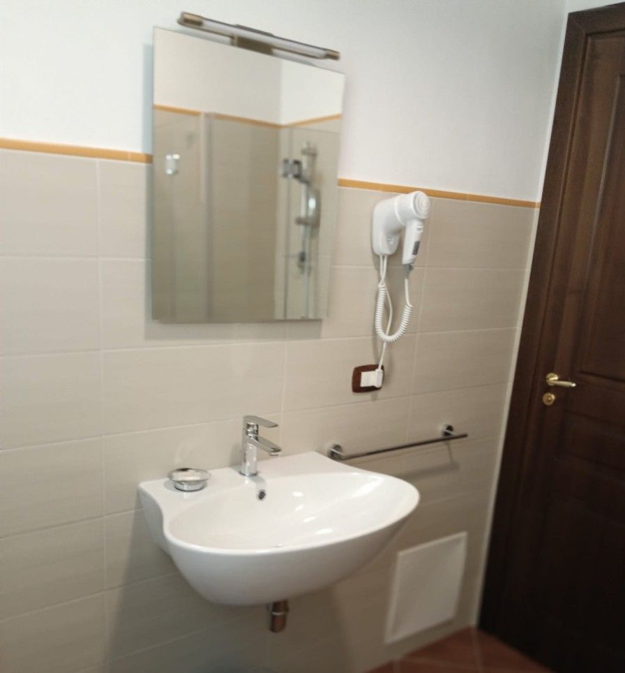 Bagno con lavandino bianco, specchio e asciugacapelli. Porta marrone sulla destra. Piastrelle beige alle pareti.