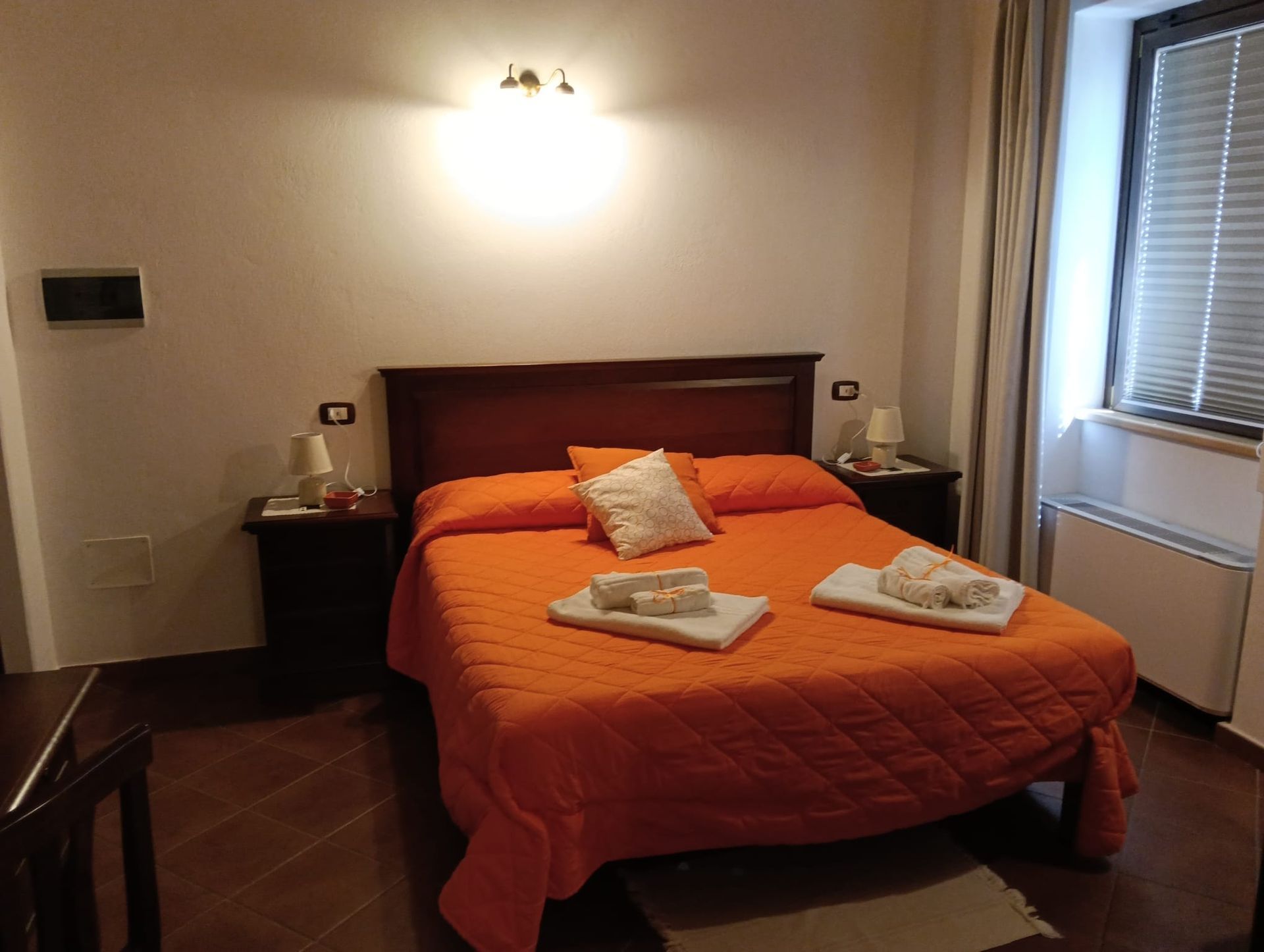 Camera da letto con copriletto arancione, due comodini e una finestra con persiane.