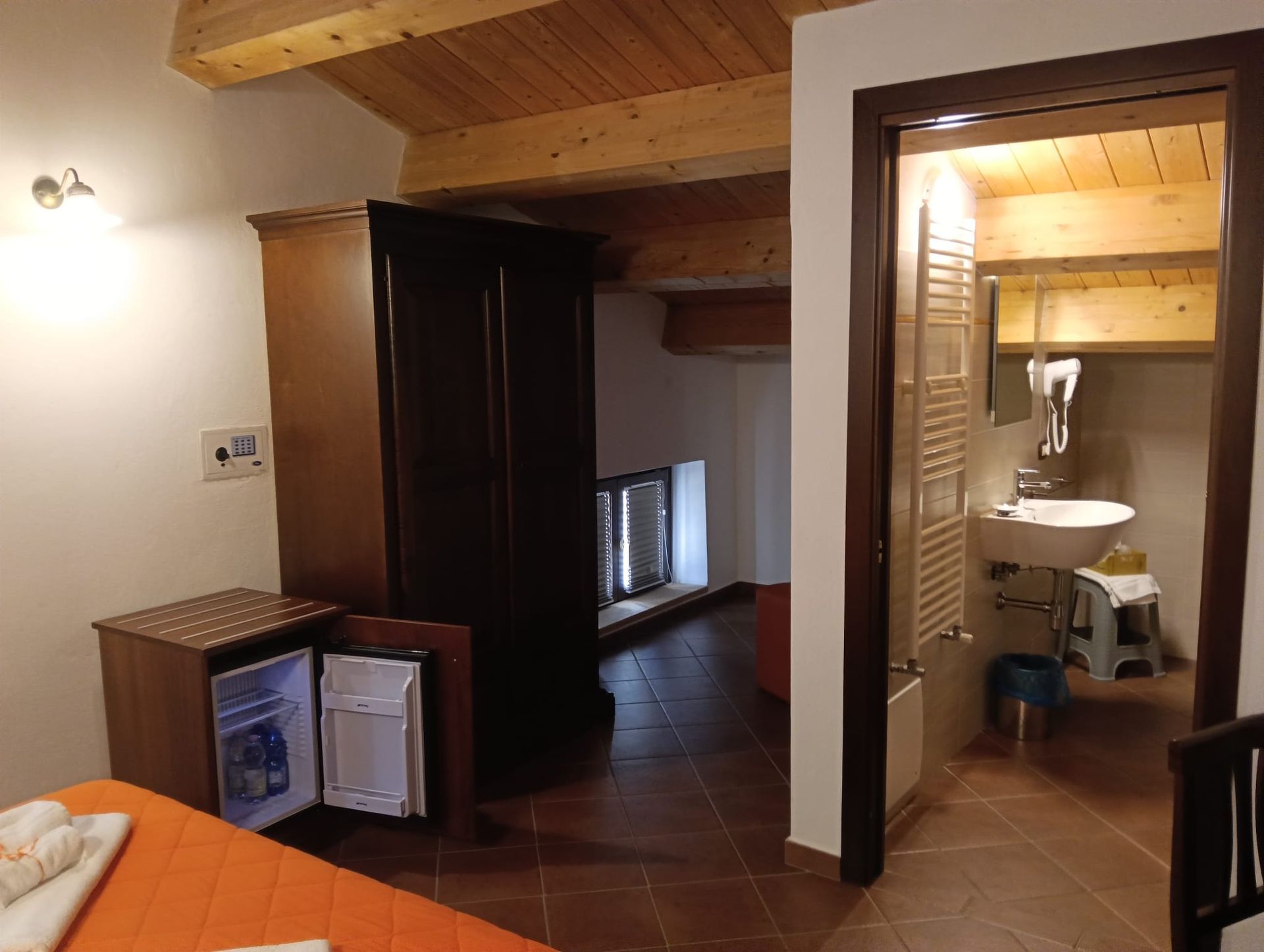 Camera d'albergo con guardaroba, frigorifero aperto, bagno a vista e travi in legno a vista sul soffitto.