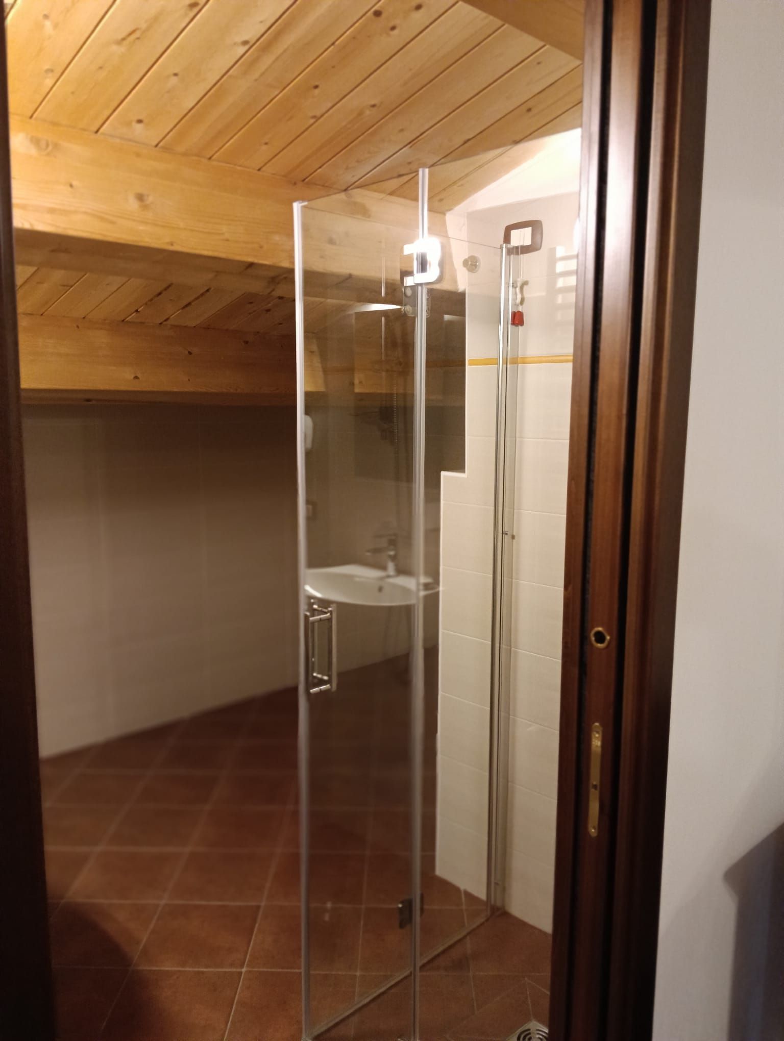 Bagno con porta doccia in vetro, soffitto in legno, pavimento piastrellato e pareti beige.
