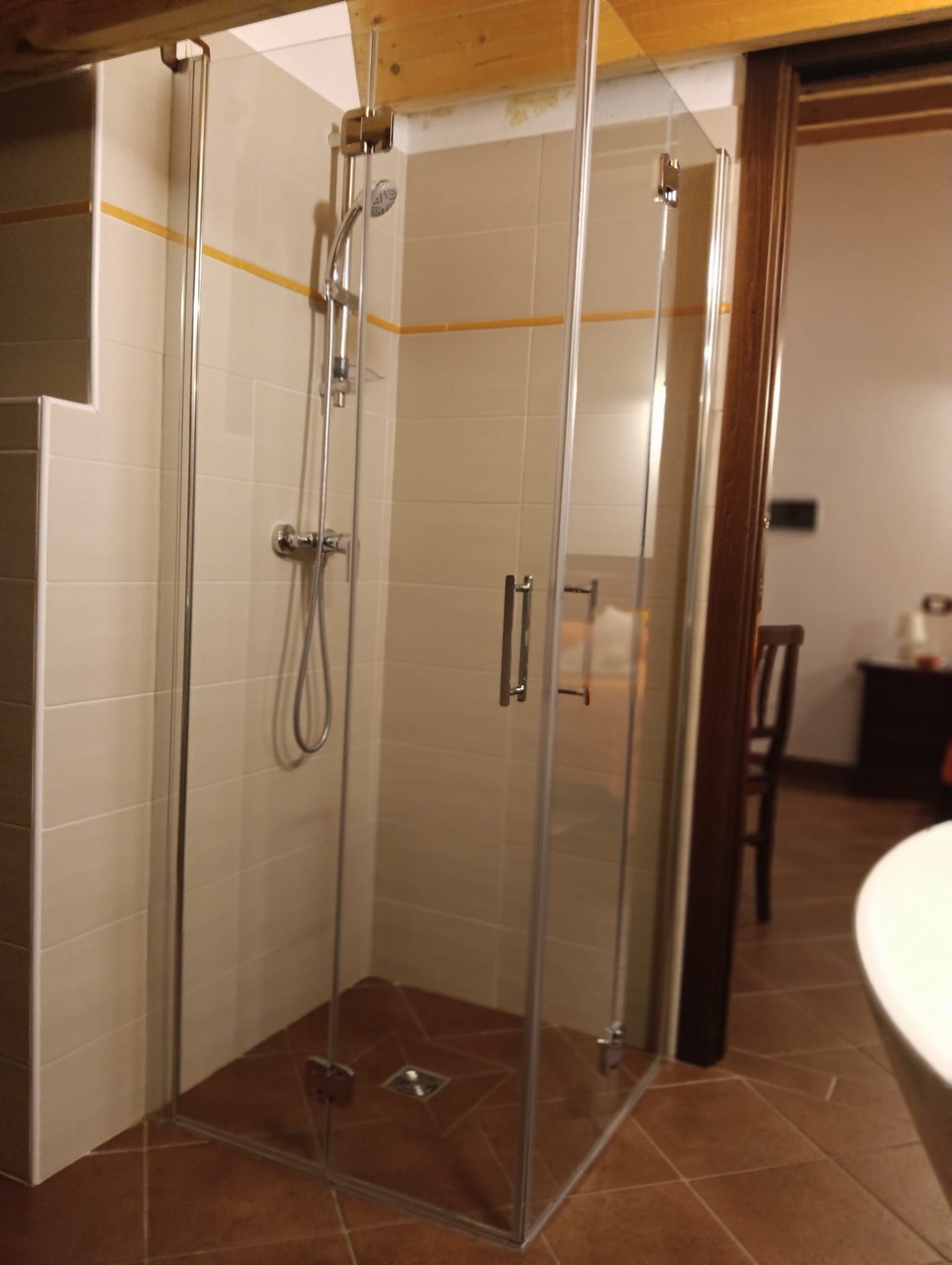 Box doccia in vetro con accessori cromati e piastrelle beige, integrato in un bagno con pavimento in cotto.